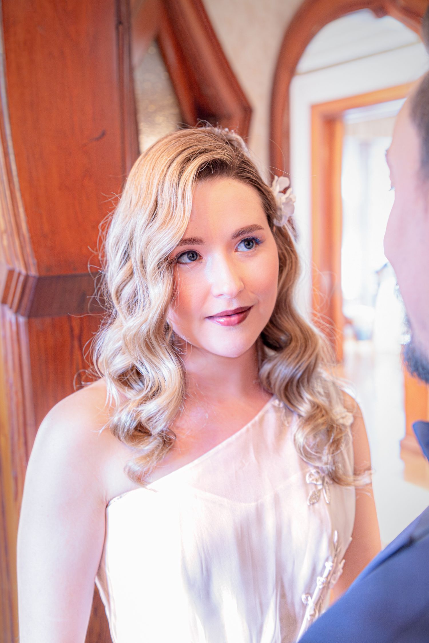 A bride in a white dress with wavy blonde hair and soft makeup smiles during her wedding day.