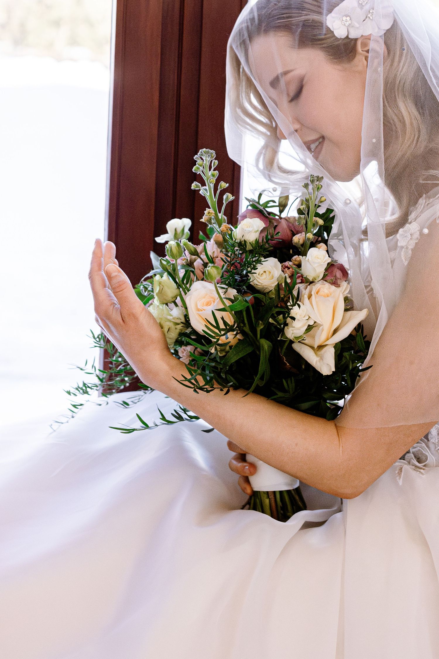 Bride in white dress admiring her wedding bouquet of roses and greenery.