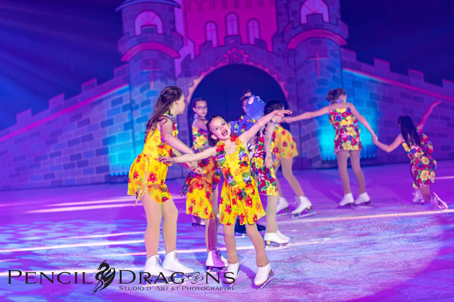 Young girls in colorful floral dresses perform an ice skating routine on a decorated stage at a holiday ice show.