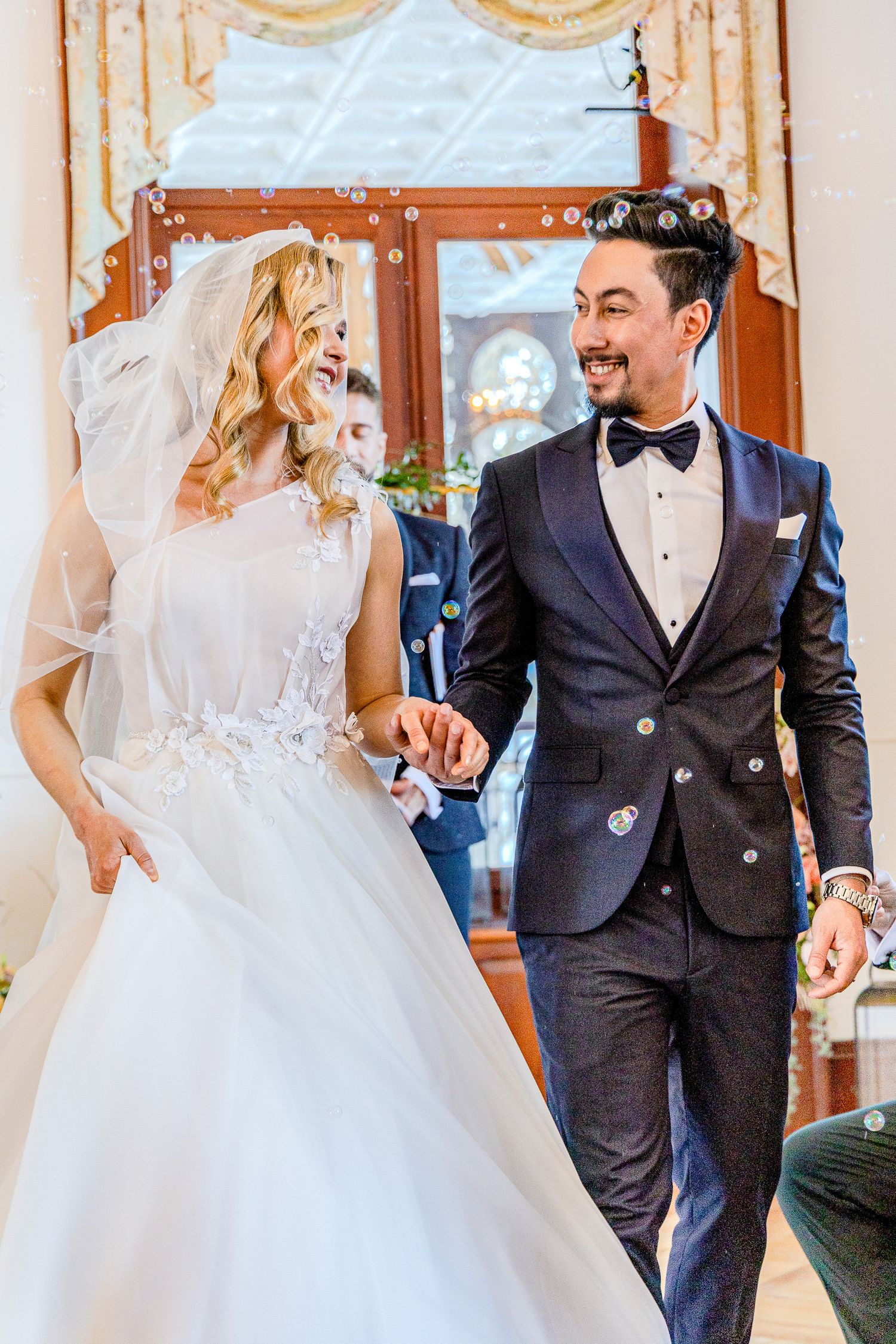 A bride and groom share a joyful moment during their wedding celebration in an elegant room with golden curtains.