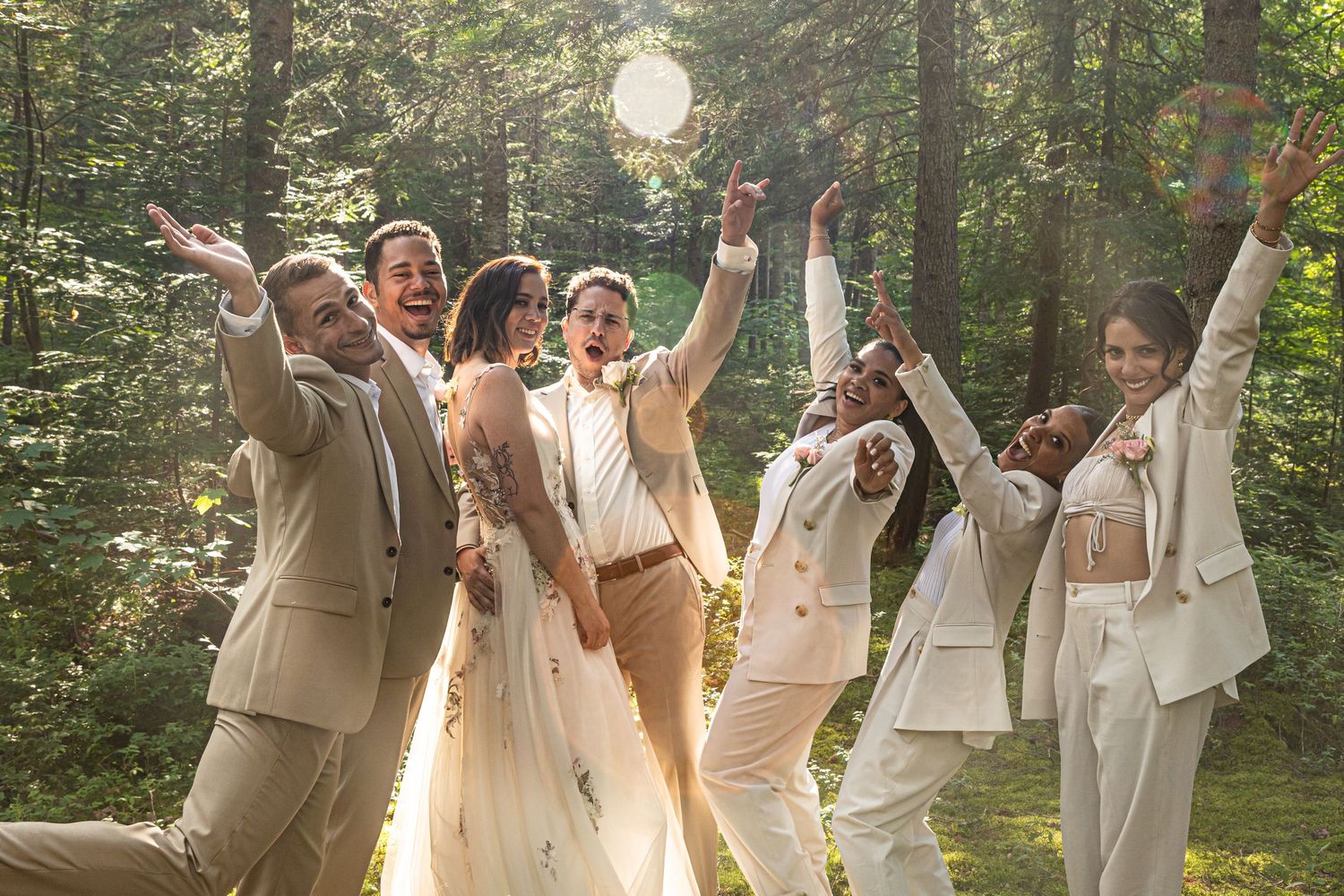 Wedding party in light colored suits celebrating joyfully in a sunlit forest setting.