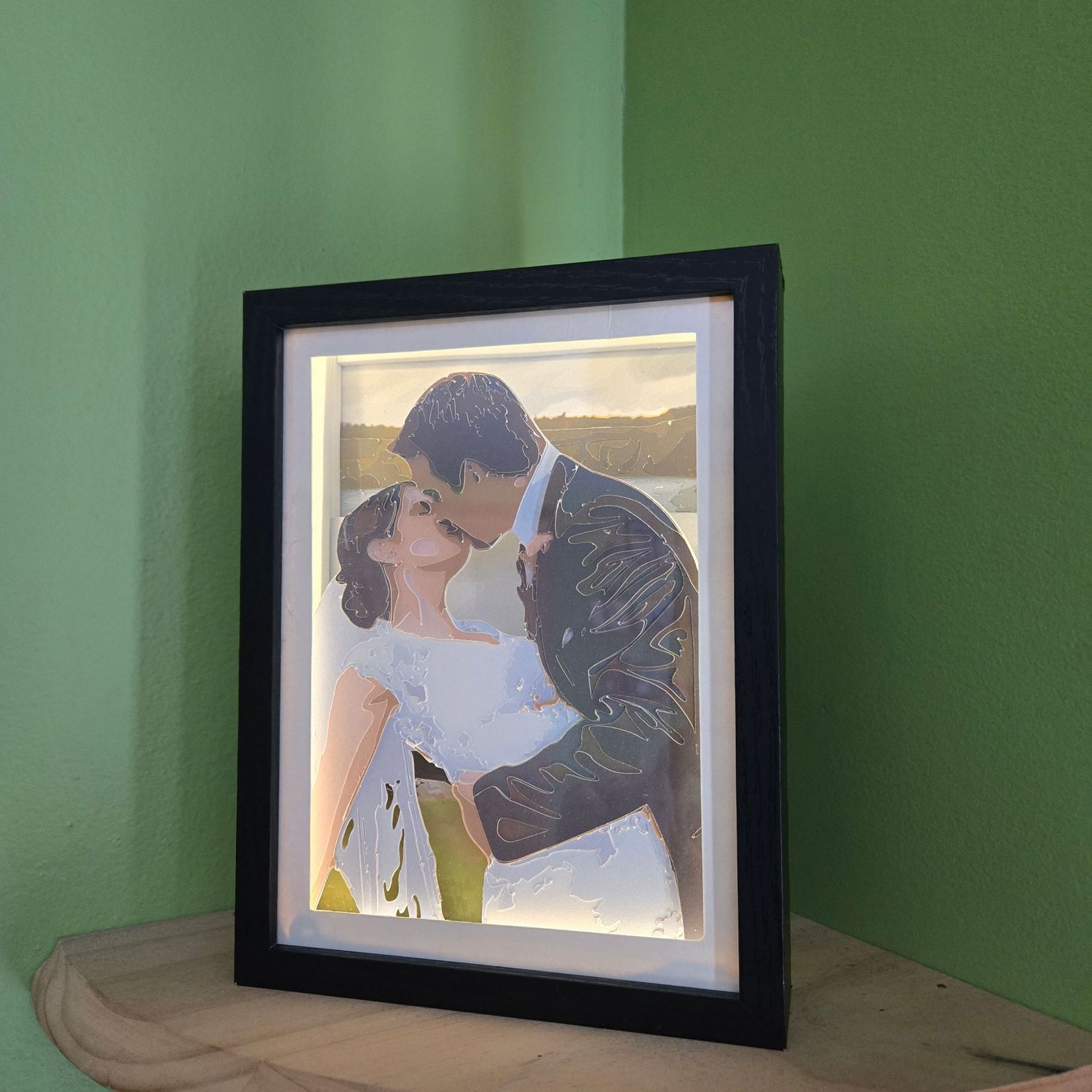 Illuminated black framed wedding portrait of a couple kissing, displayed on a wooden surface against a green wall.