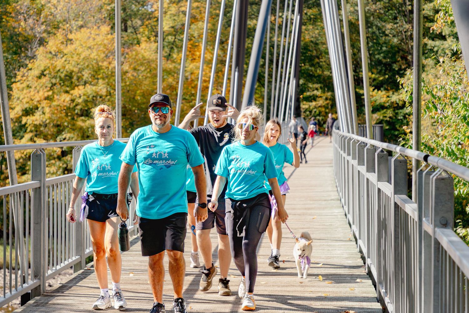 Group of people in teal shirts walking across a suspension bridge during a charity walk event on a sunny fall day.