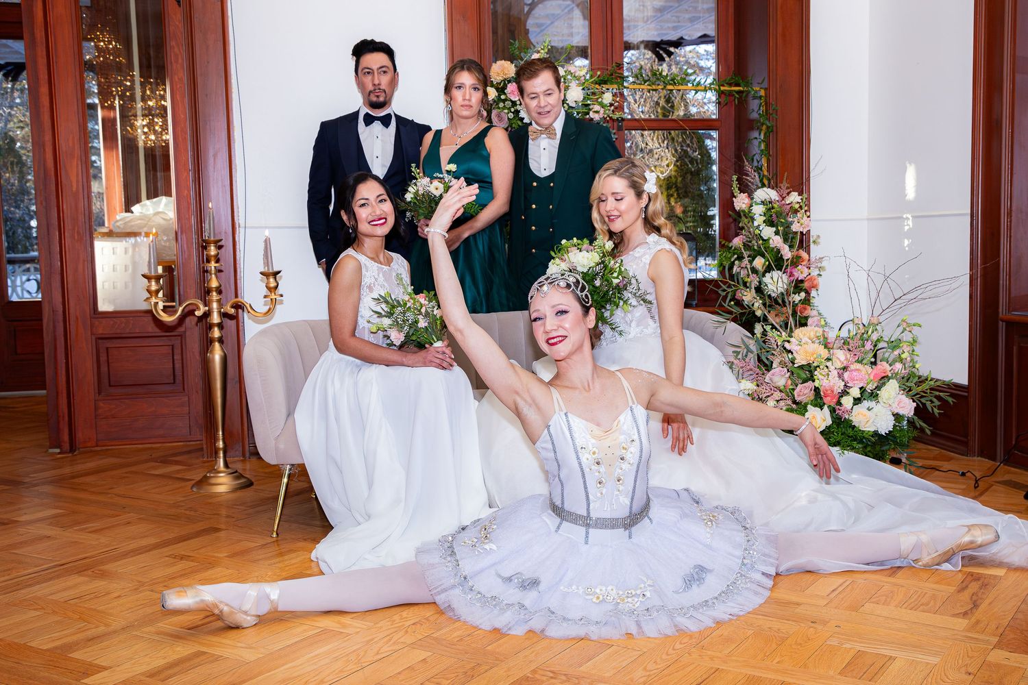 Wedding party in formal attire poses elegantly on hardwood floor with floral decorations and candelabras in background.