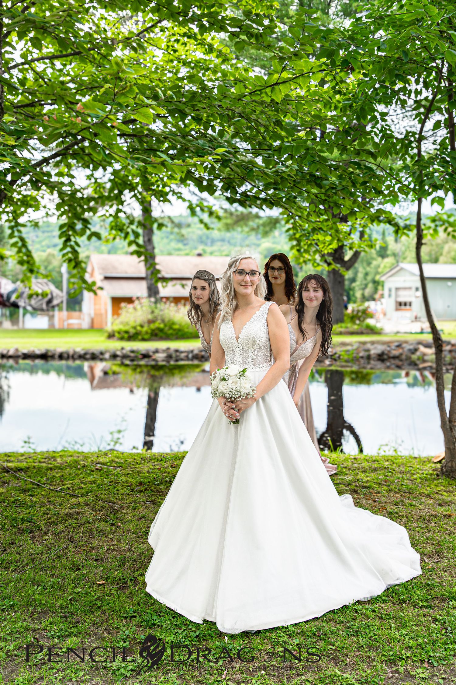 Wedding party in white dresses poses under green trees by a lakeside.