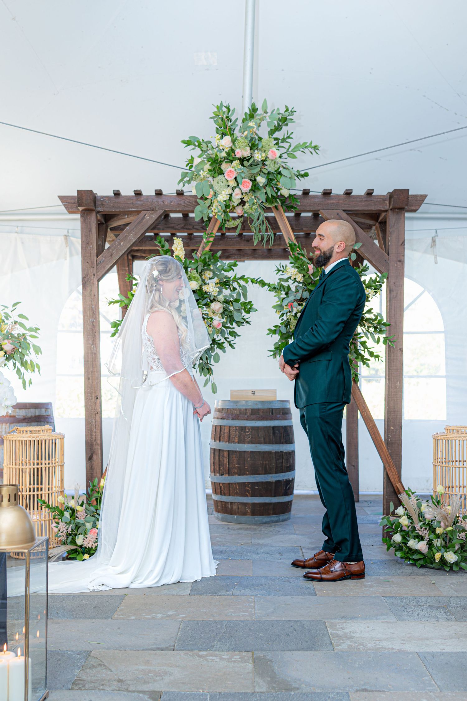 Wedding ceremony setup under wooden pergola with wine barrels and floral decorations.