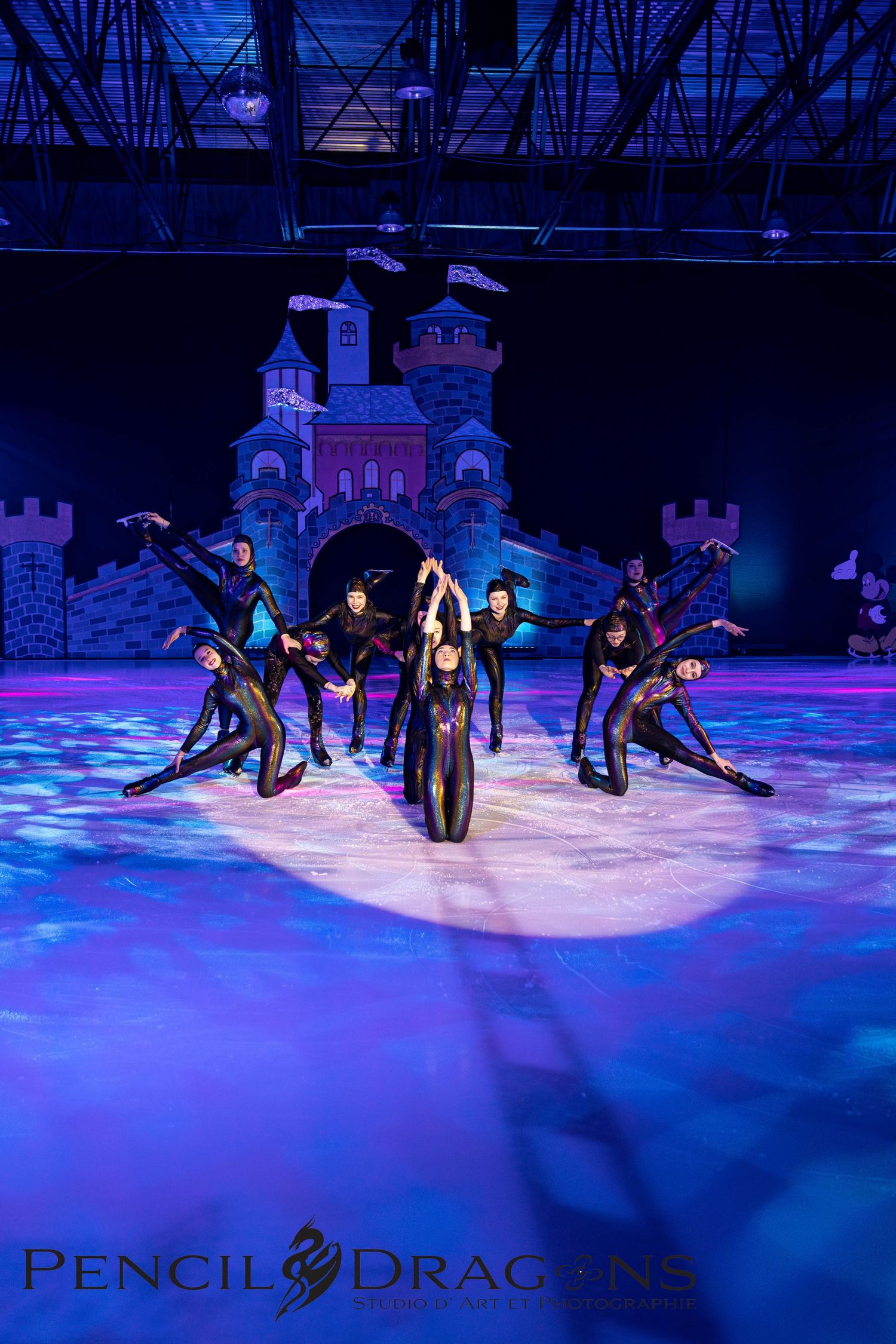 Ice skaters in black costumes perform a synchronized routine on a rink with a castle backdrop and blue stage lighting.