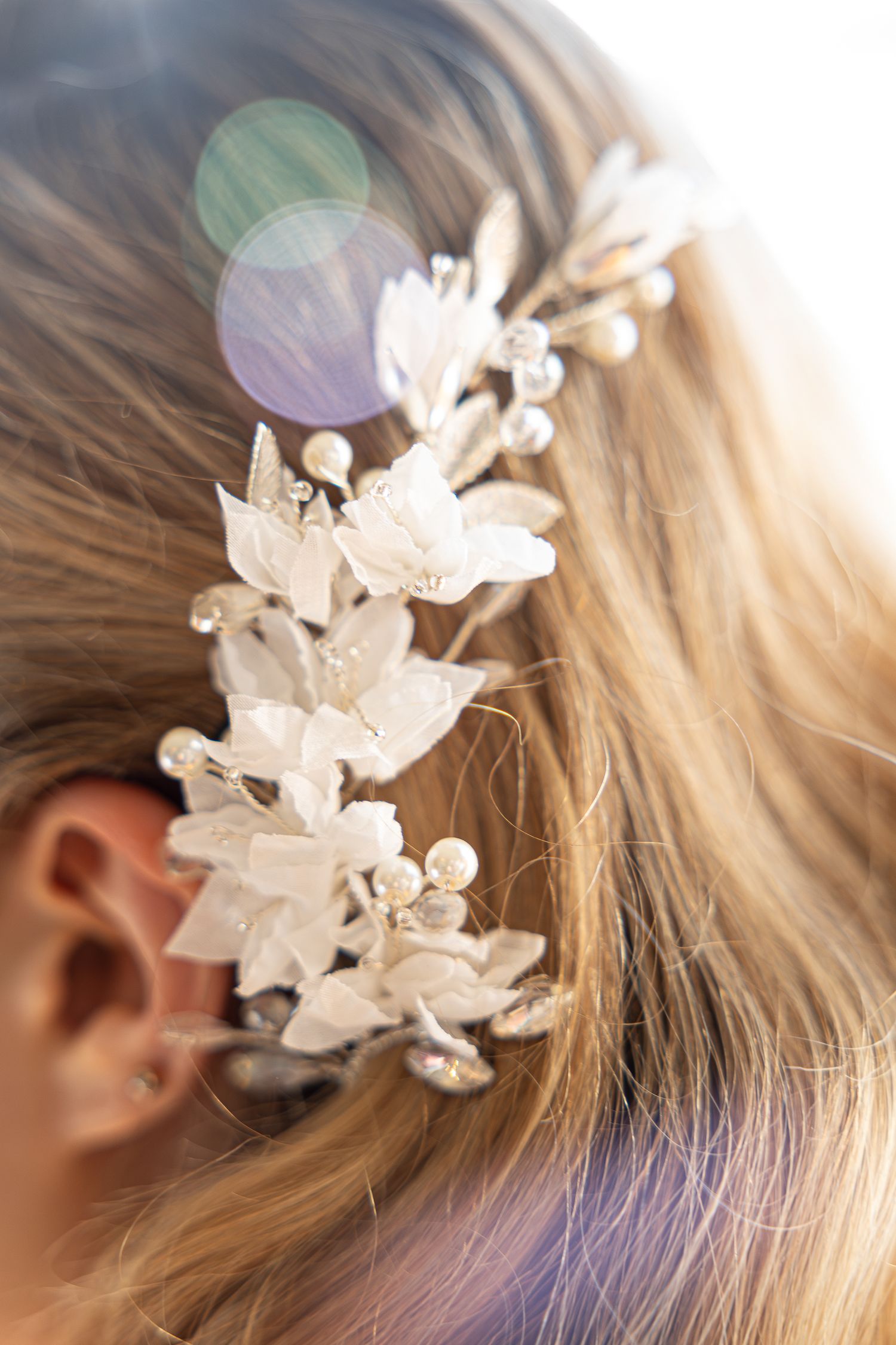 A delicate white floral hair accessory with pearls adorns blonde hair in soft lighting.