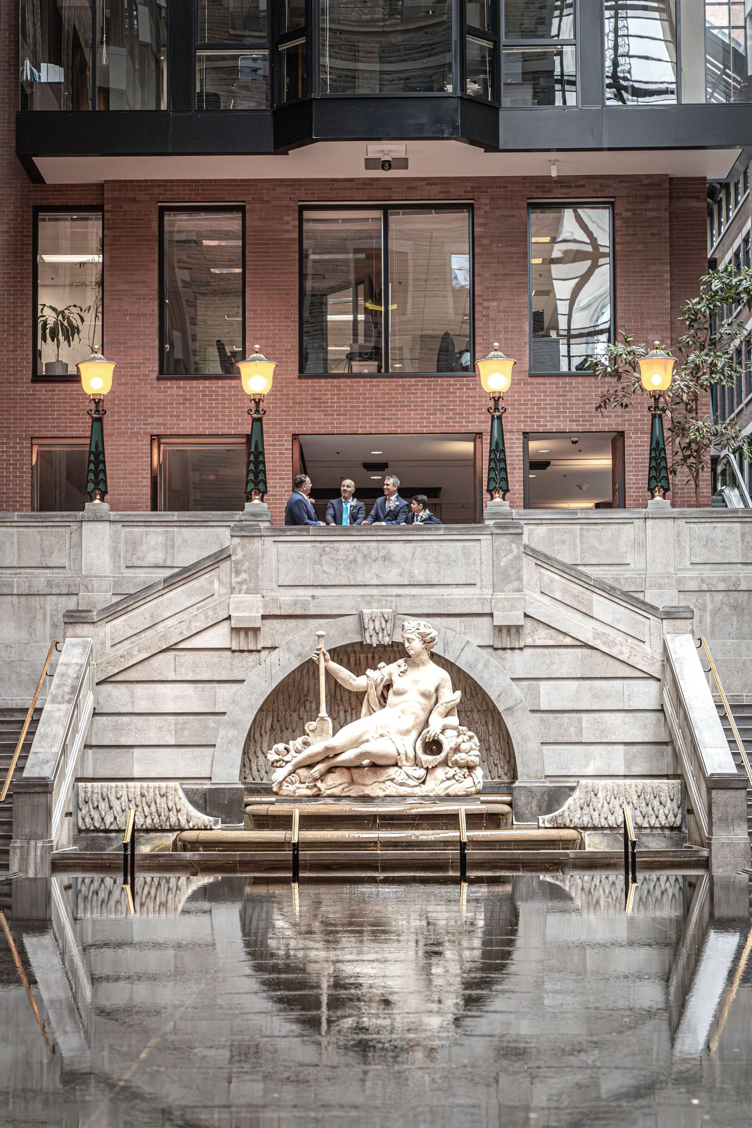 Modern brick building with illuminated wall sconces and decorative stone archway featuring classical relief sculpture reflected in water.