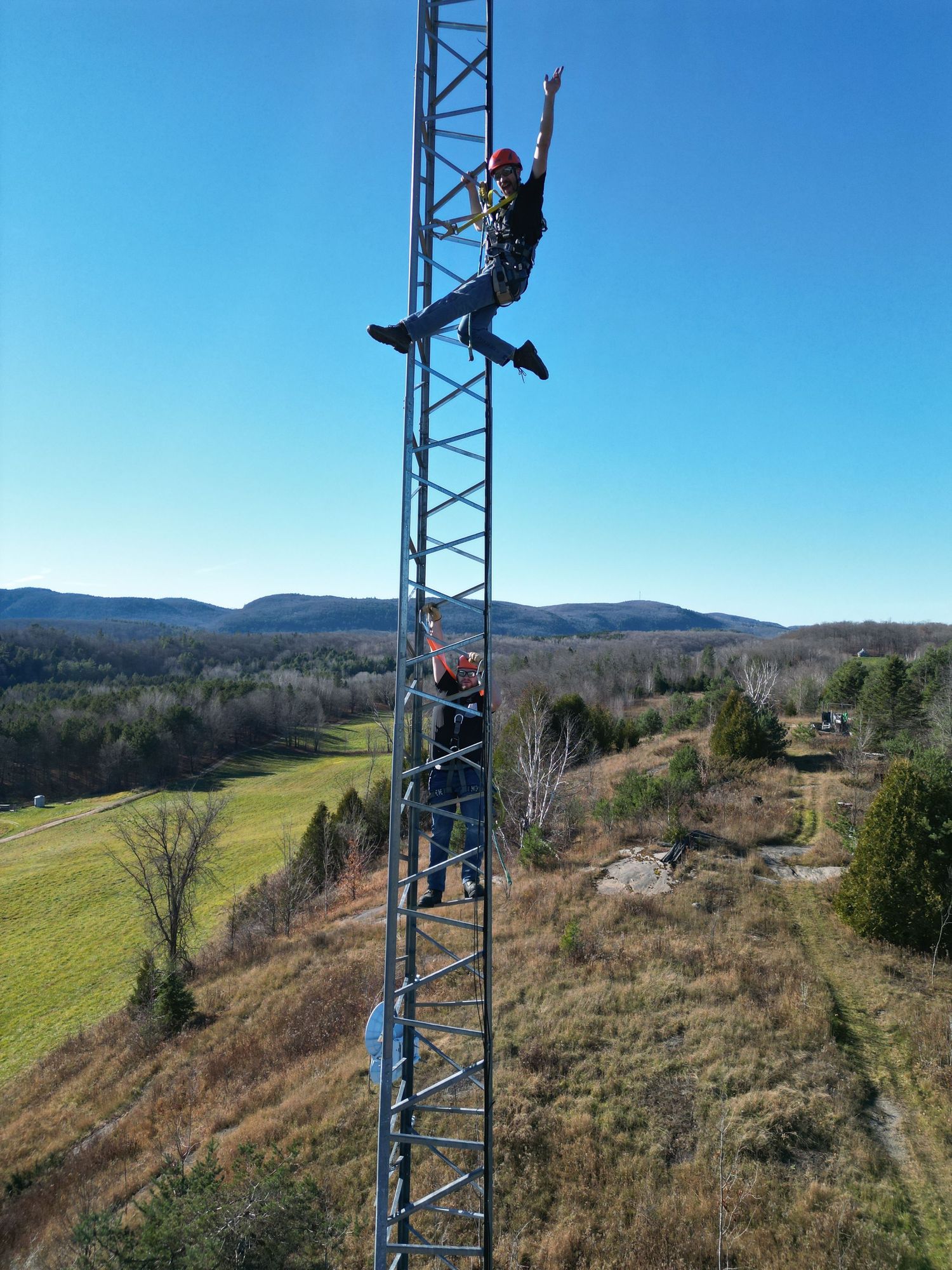 Two workers climbing a tall metal communications tower with scenic rural landscape and blue sky backdrop.