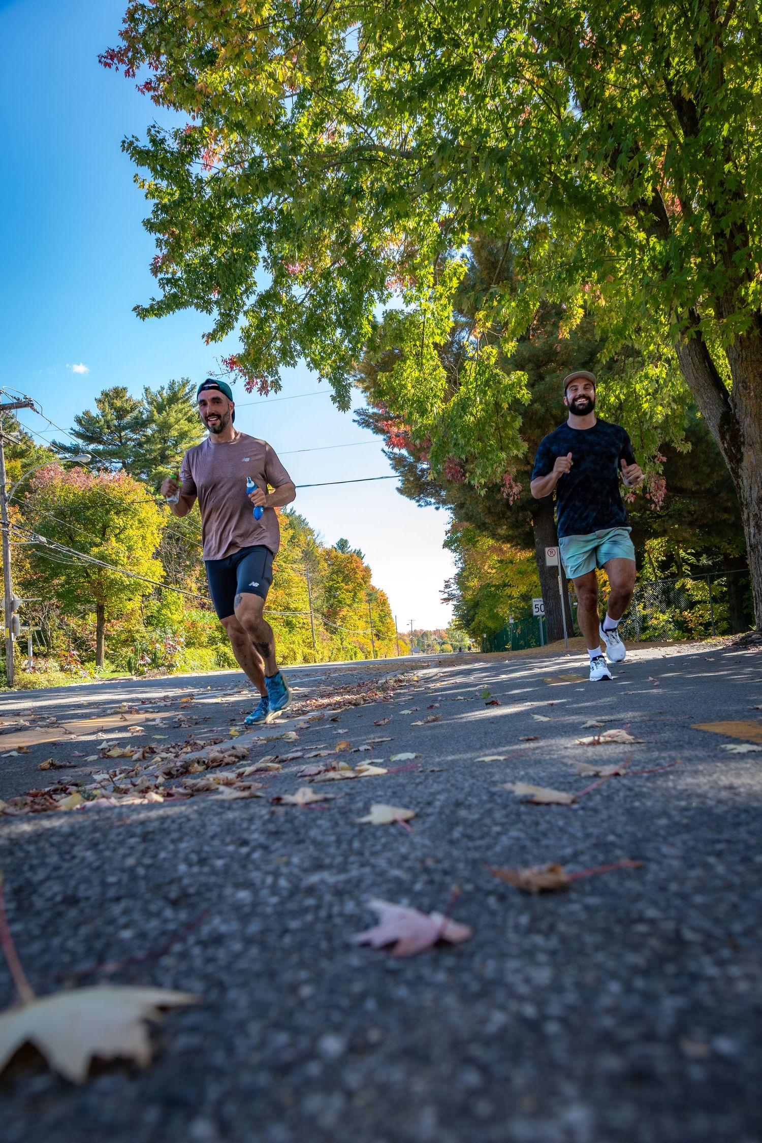 Two men jogging on a tree-lined road in autumn, with colorful foliage and fallen leaves under a bright blue sky.