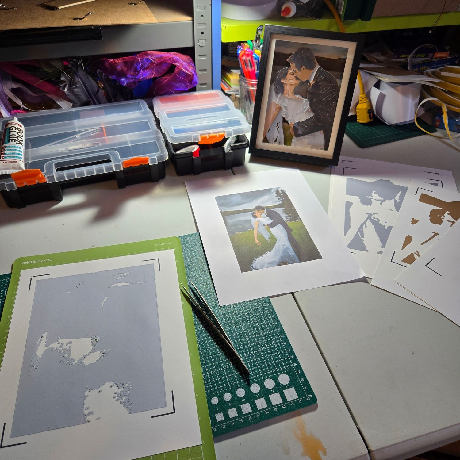Craft workspace with wedding photo, cut paper stencils, cutting mat, craft knife, and organizer boxes on a desk.