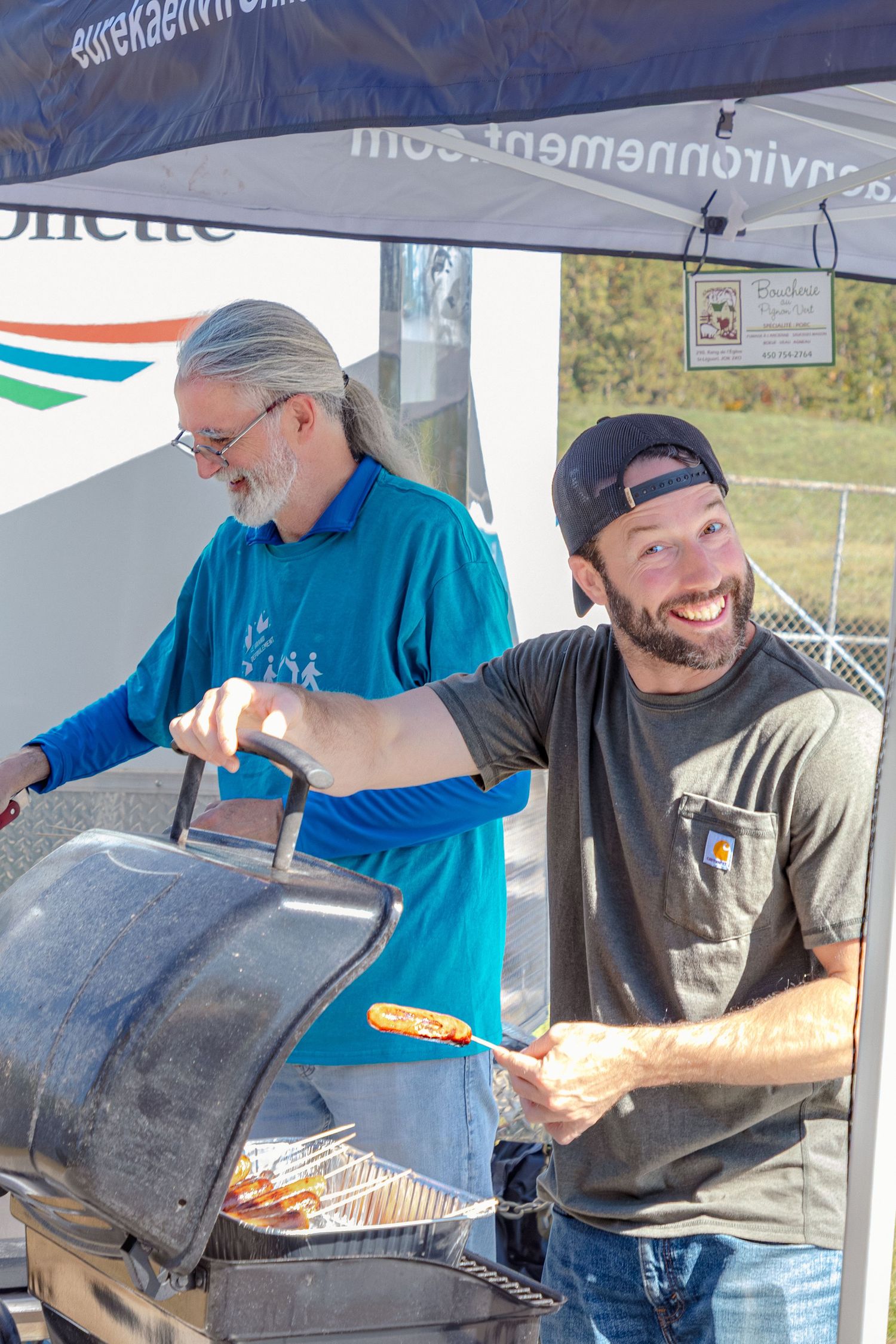 Two men grilling hot dogs and food outdoors under a canopy tent at a community barbecue event.