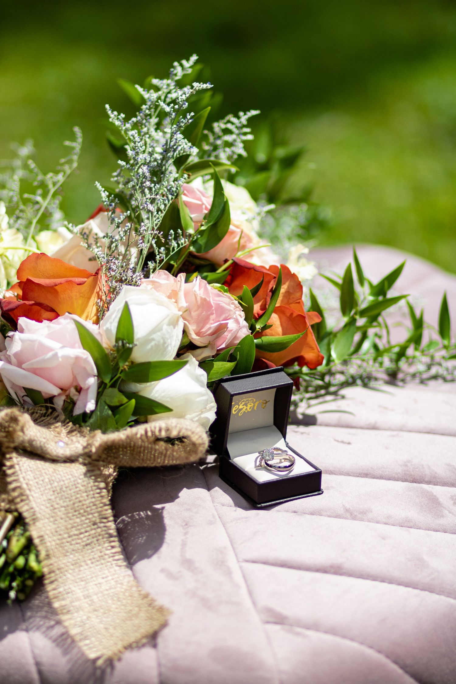 Wedding bouquet with pink and coral roses alongside a ring box on quilted fabric.