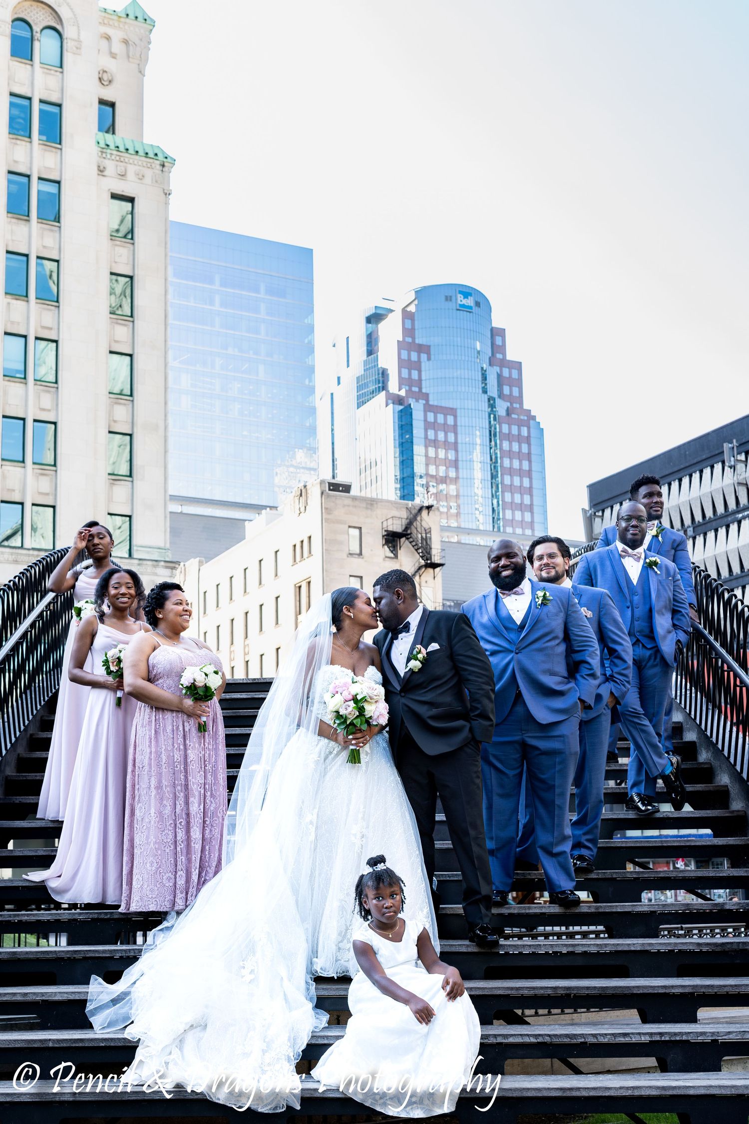 A wedding party posed on grand outdoor steps with city buildings in the background.
