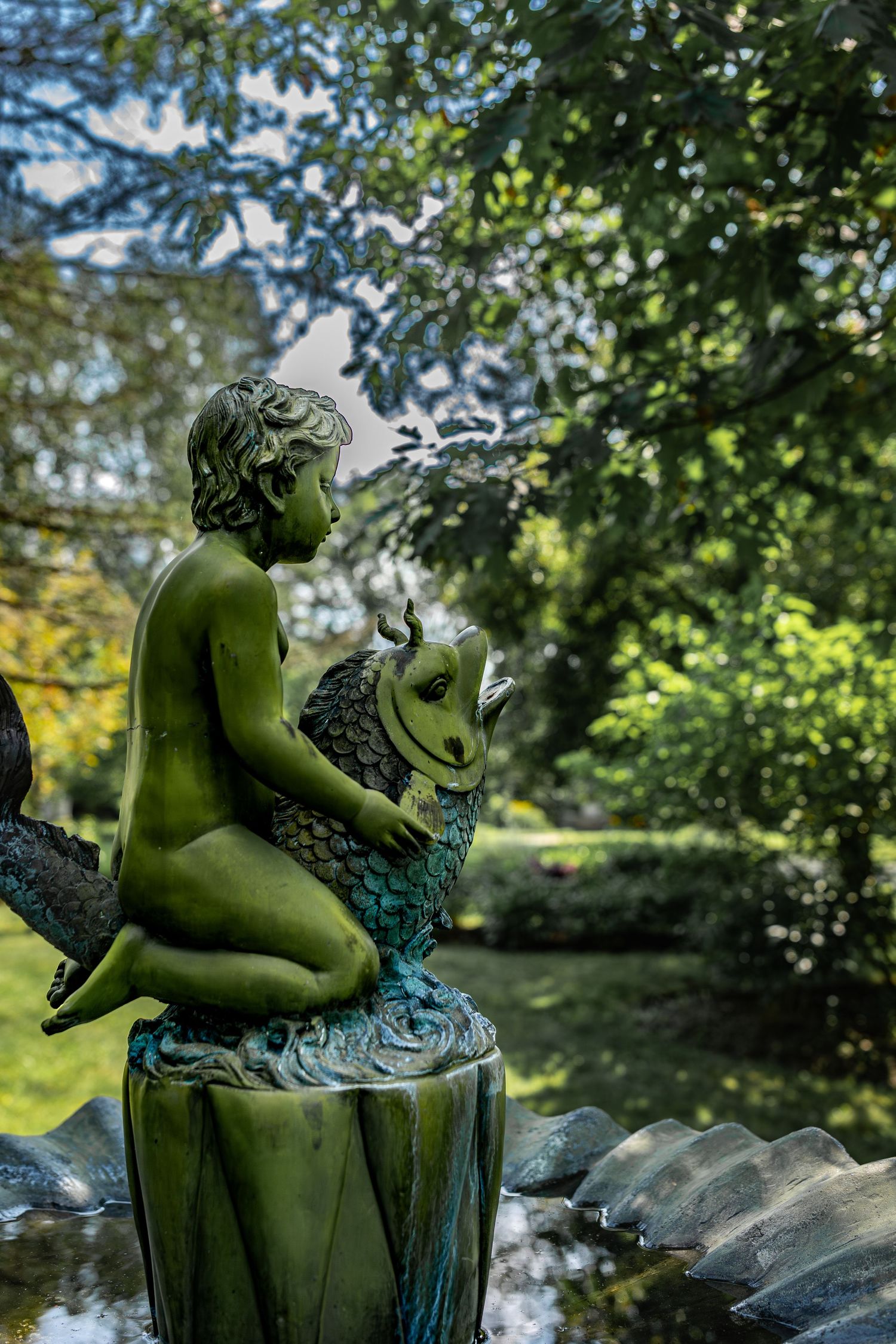 Garden statue of a cherub riding a fish ornament in verdigris patina, surrounded by lush green foliage.