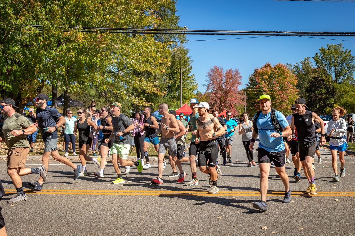 Large group of runners starting a road race on a sunny autumn day with colorful trees lining the street.