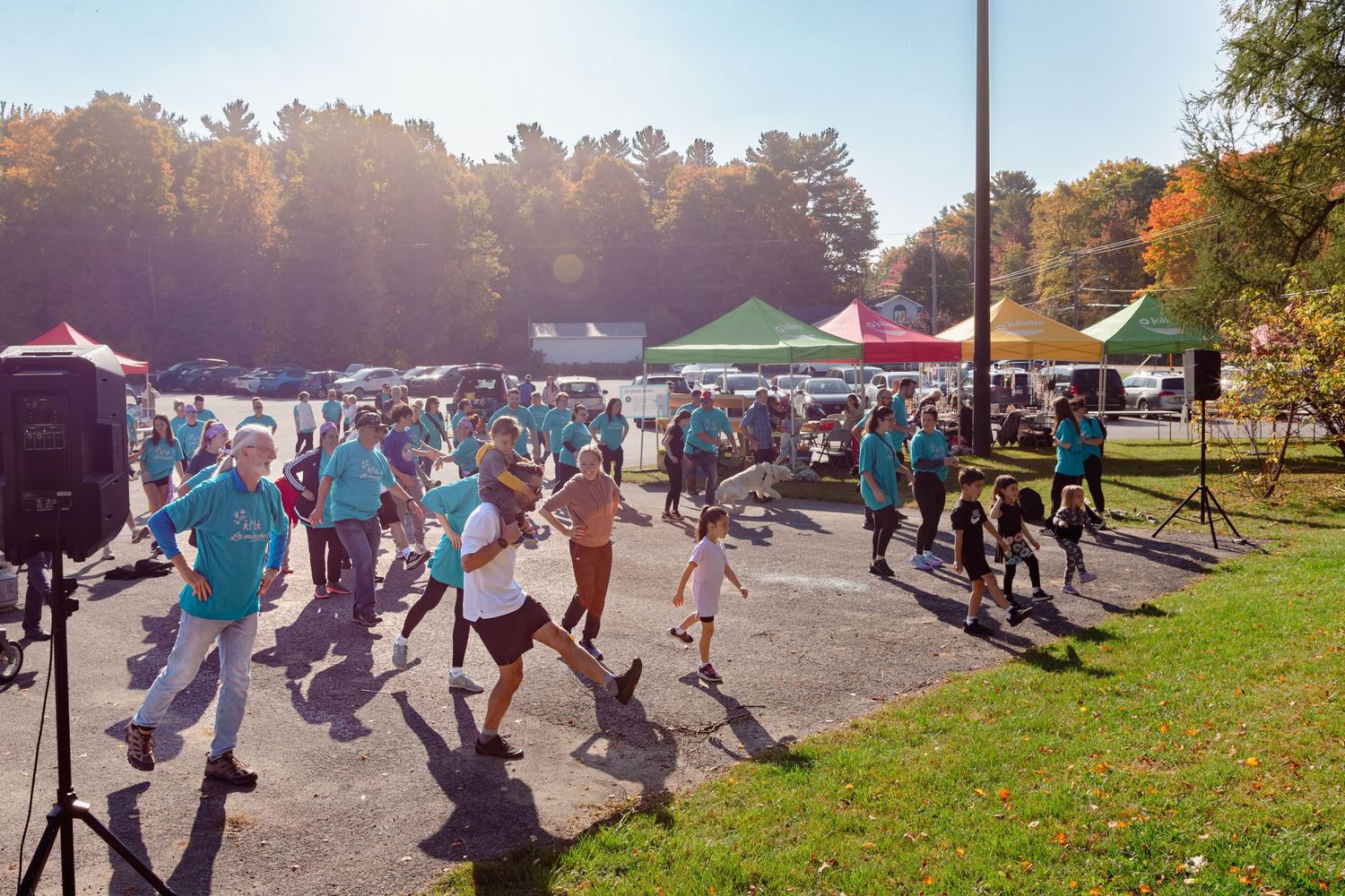 Crowd of people in teal shirts walking at an outdoor charity walk event with colorful tents on a sunny autumn day.