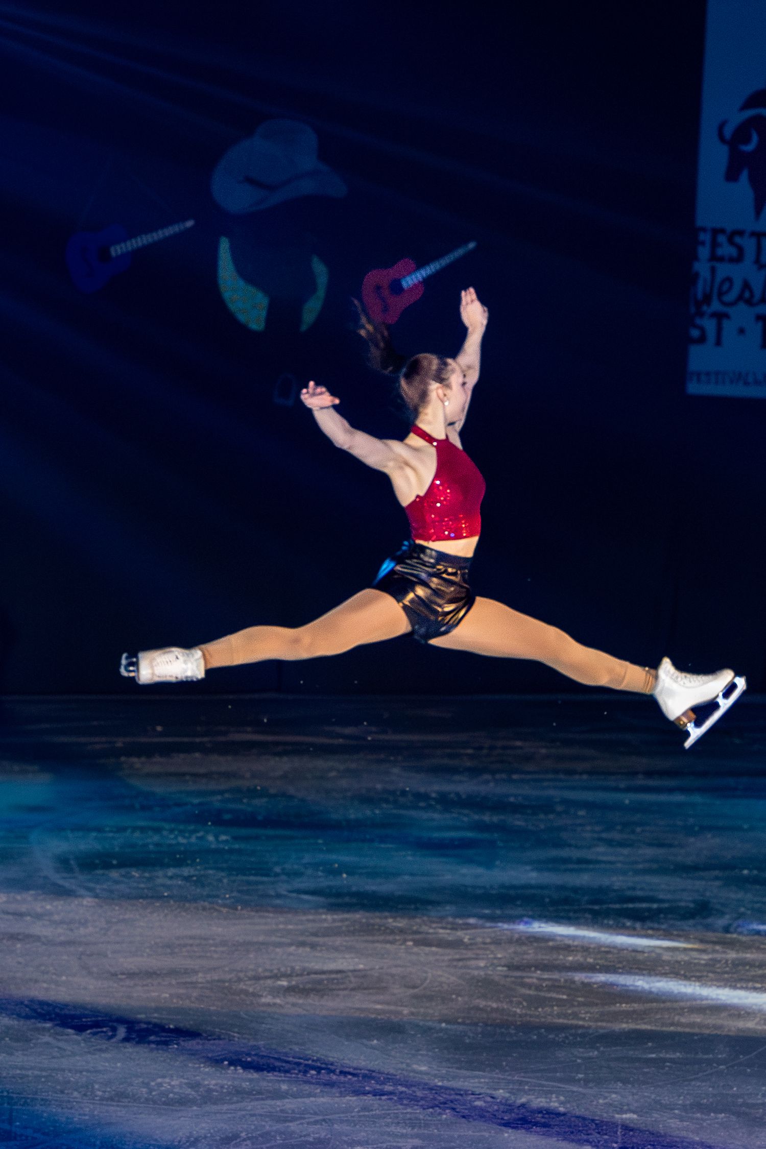 Female figure skater performing a spectacular split jump in mid-air wearing a red costume and white ice skates on an indoor rink.
