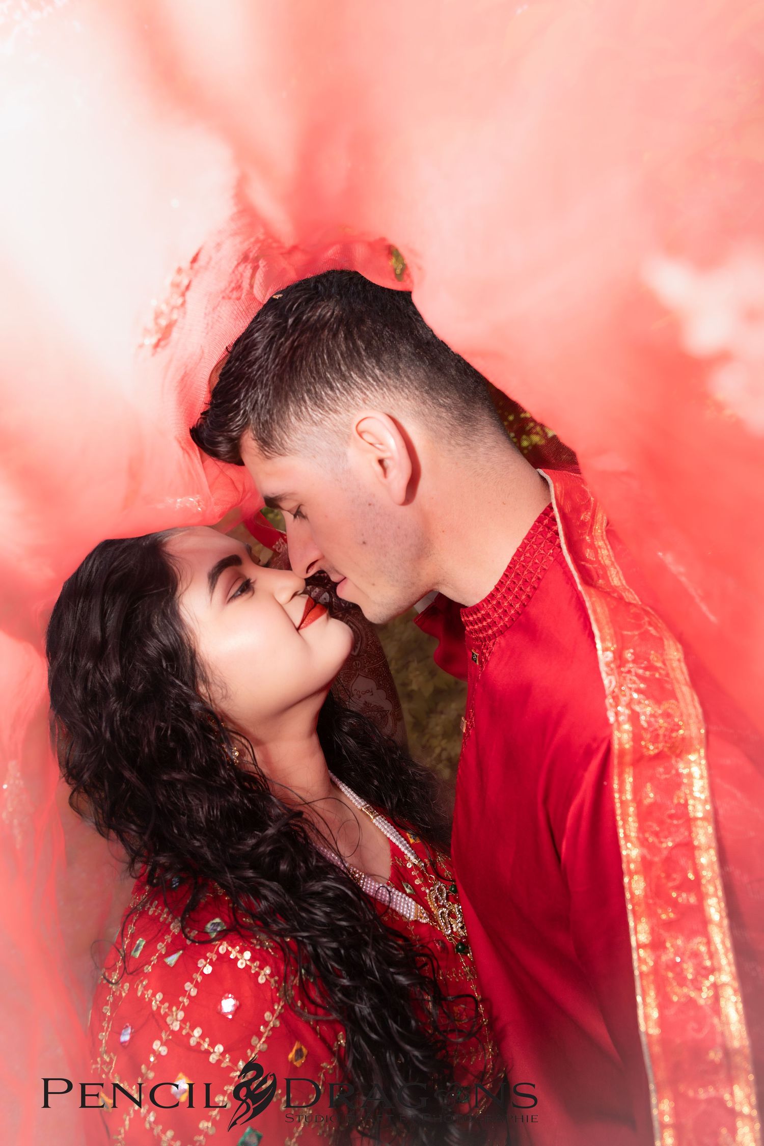 Couple in red traditional attire sharing romantic moments against a coral-colored background.