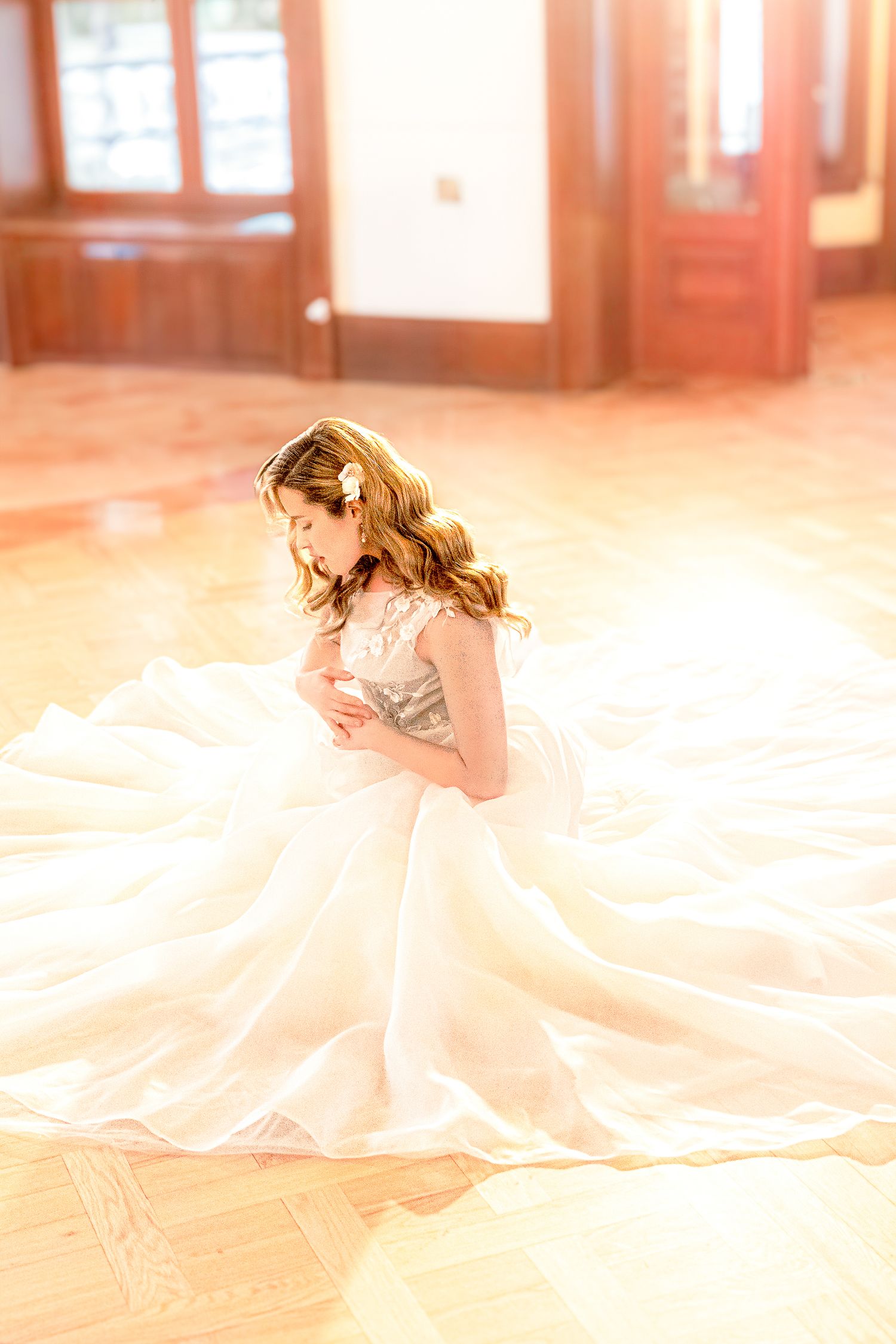 A bride in a flowing white gown lies gracefully on a sunlit wooden floor in an elegant indoor setting.