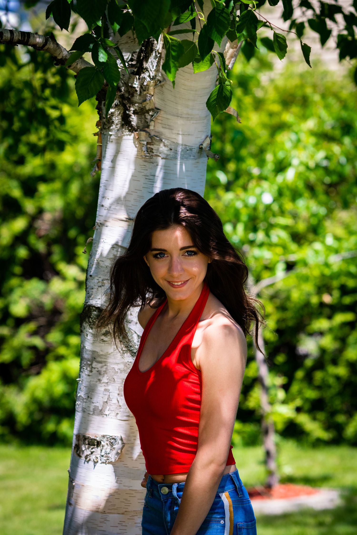 Woman in red tank top poses playfully near white birch trees in a summer garden setting.