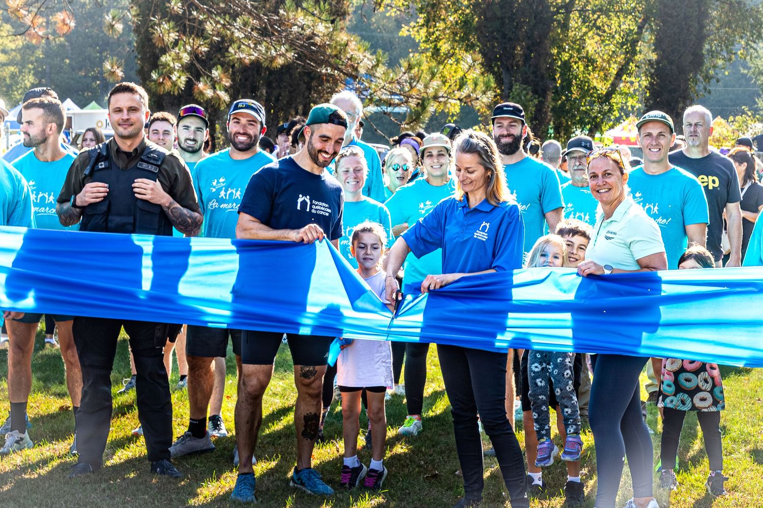Group of people in teal shirts cutting a blue ribbon at an outdoor charity run event on a sunny day.