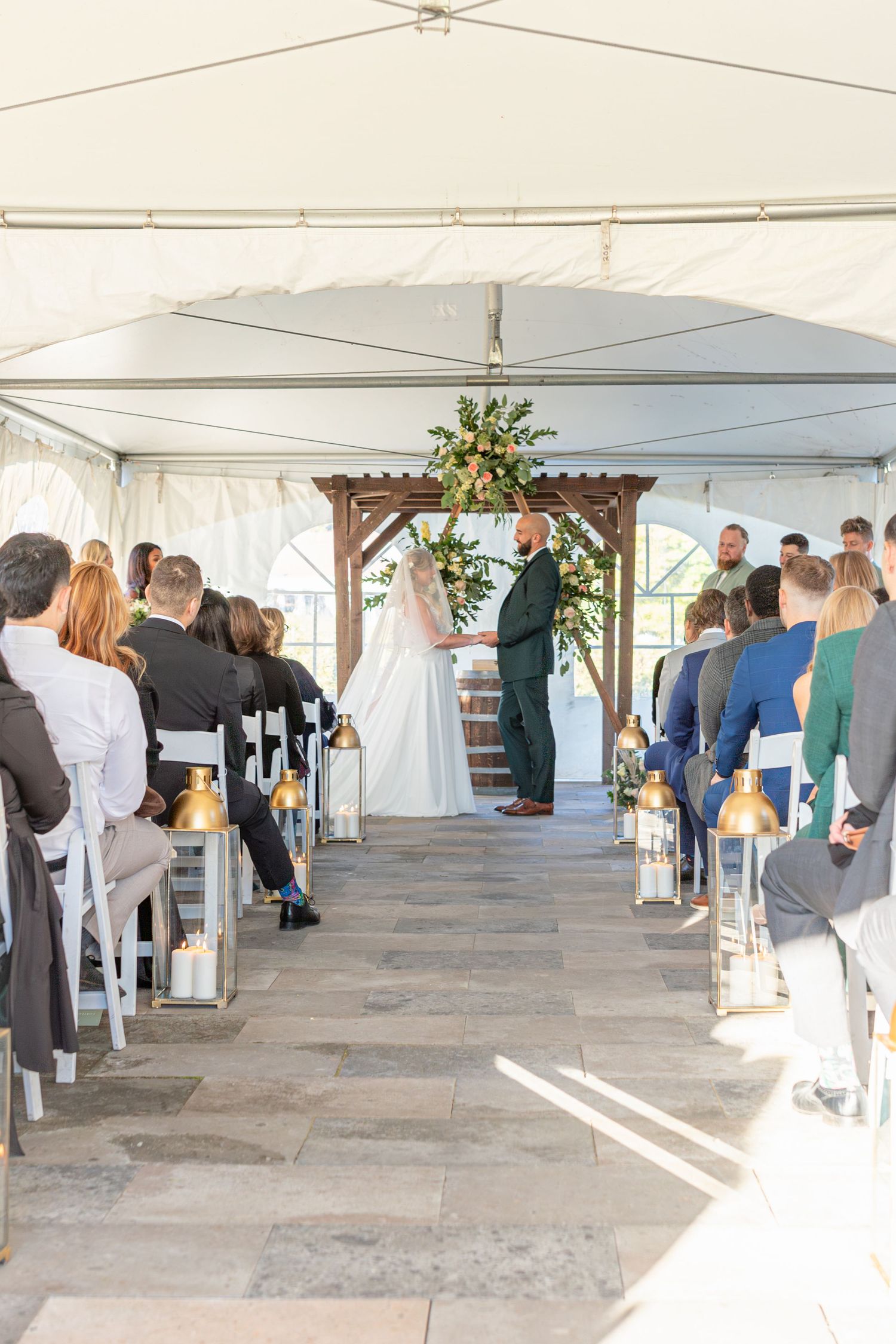 Wedding ceremony in a white tent with guests seated watching the ceremony under floral decorations.