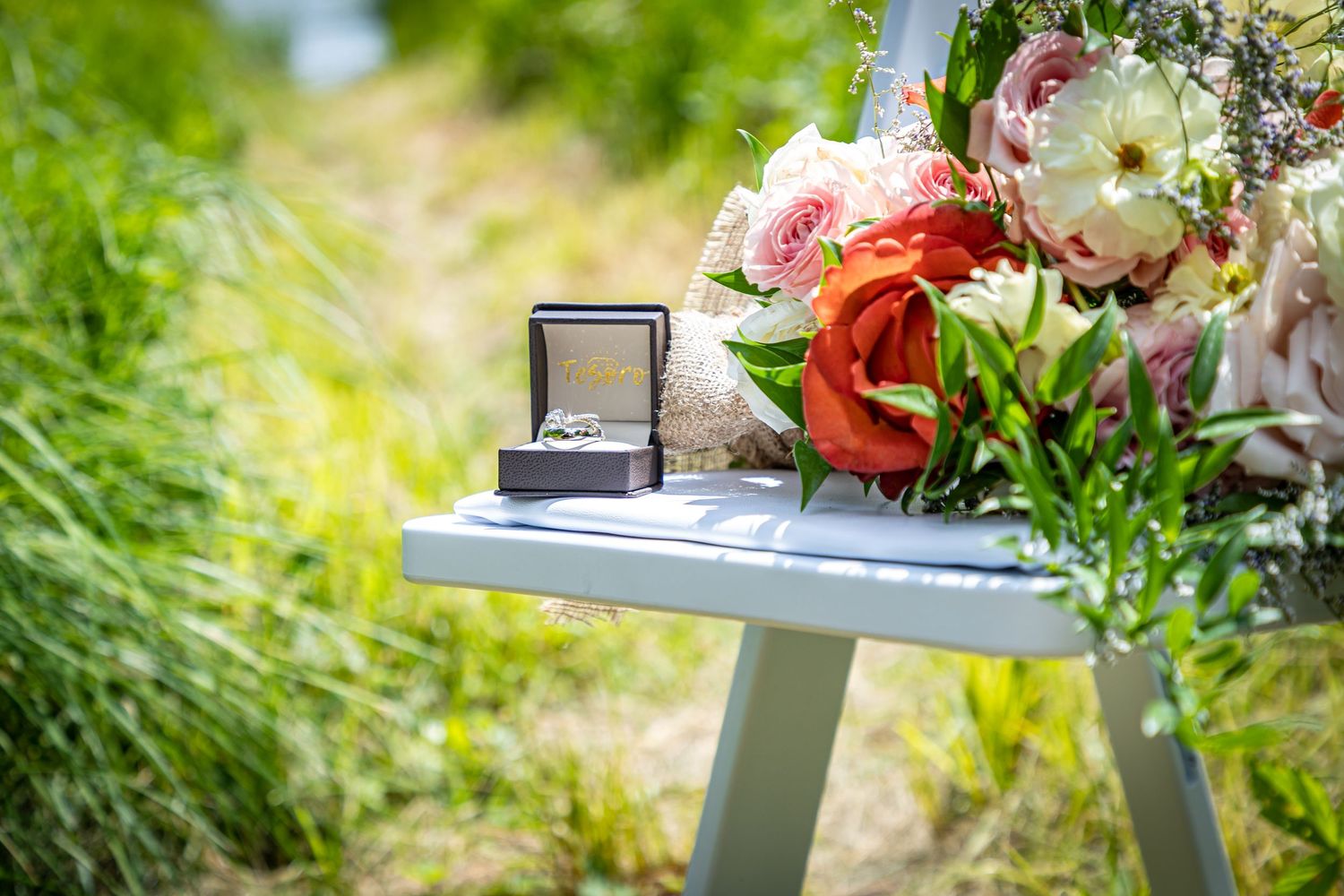 Wedding rings in a box displayed with pink and white floral arrangements on a small table outdoors.