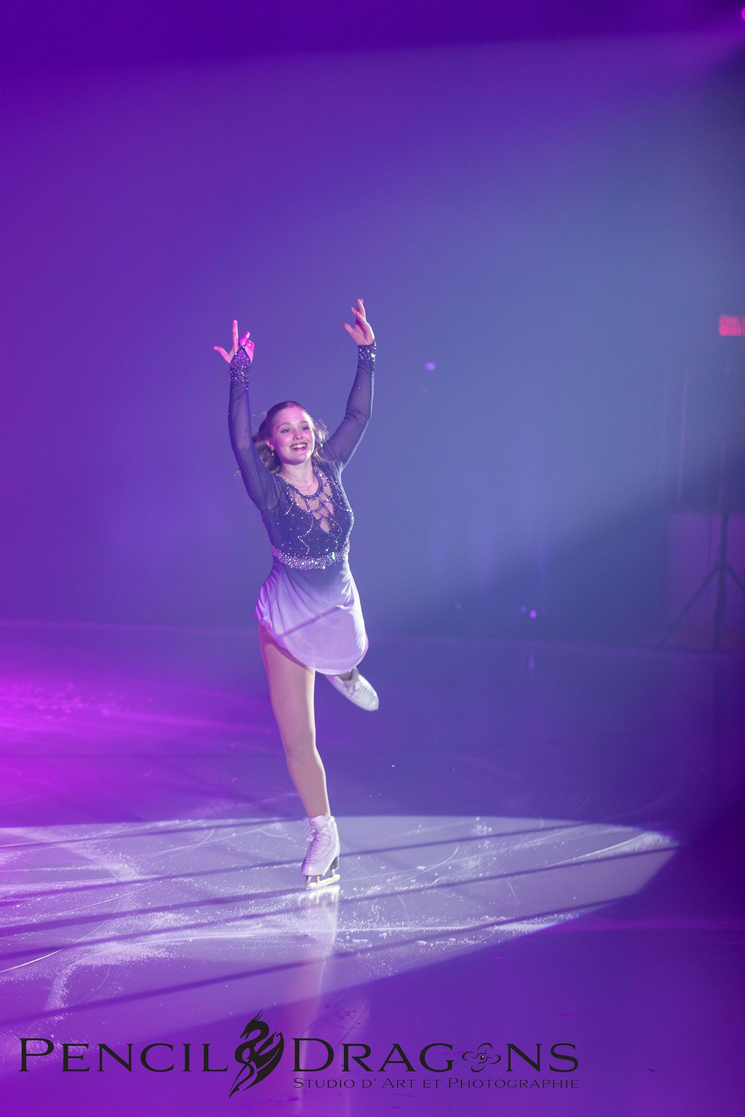 Young female figure skater performs gracefully on ice in a purple-lit rink, wearing a black and white dress with arms raised.