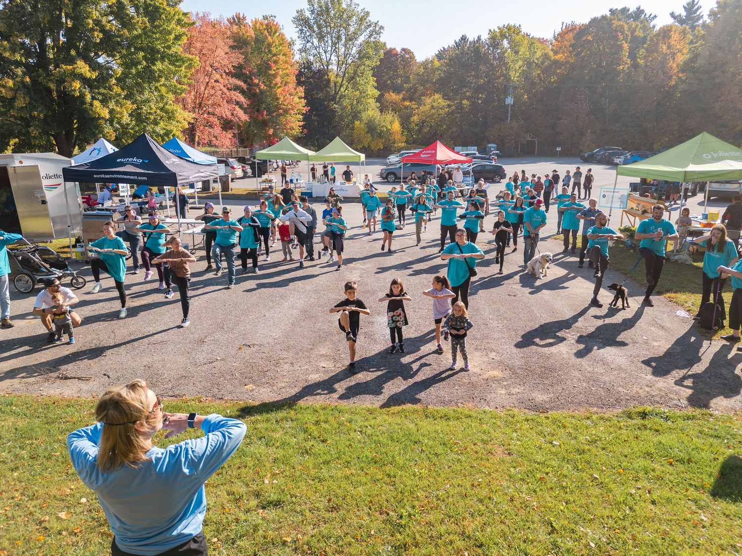 Large group in teal shirts doing outdoor fitness event in autumn park with colorful tents and fall foliage.
