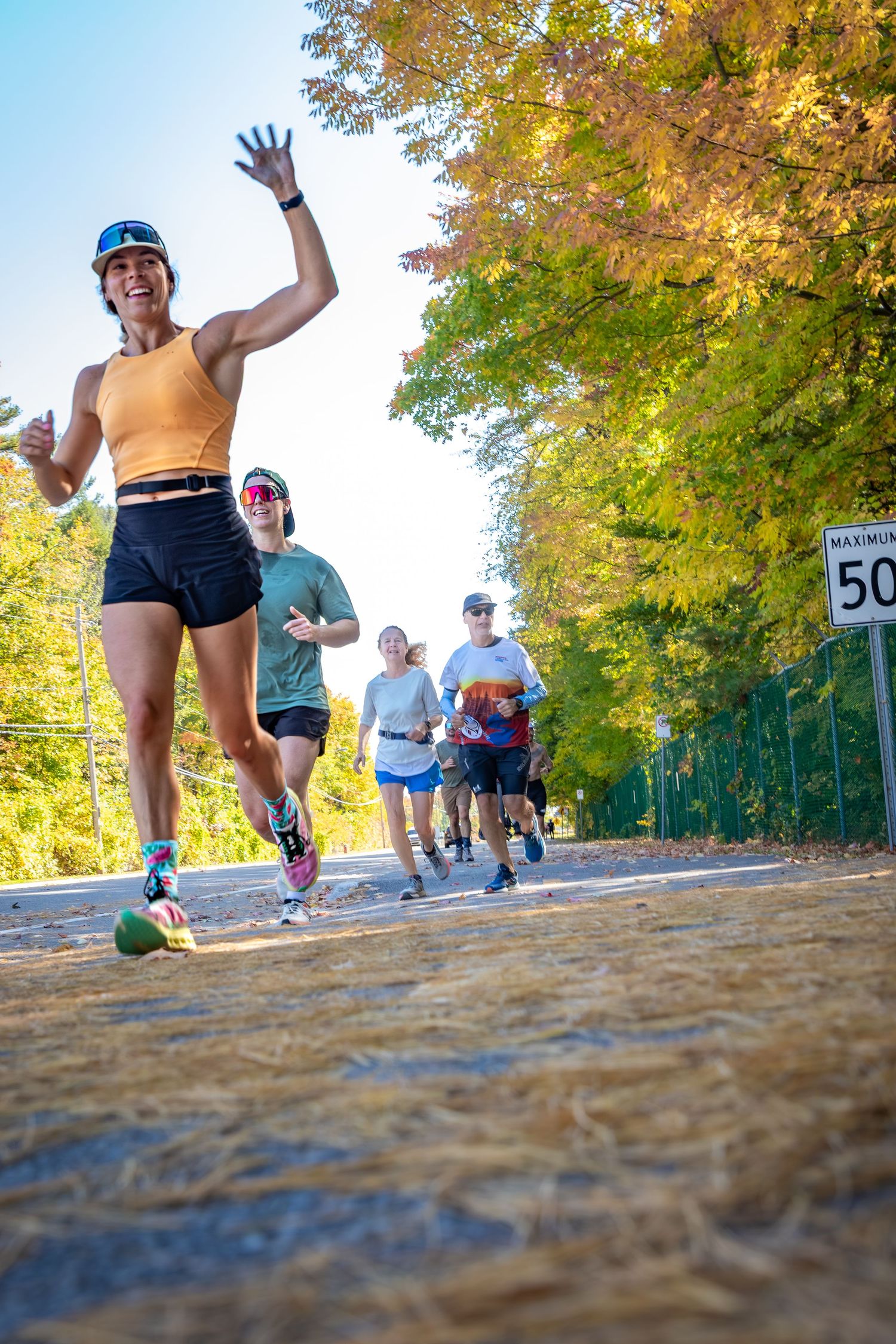 Runners race down a tree-lined road in fall foliage, with a smiling woman in orange top waving at the camera.