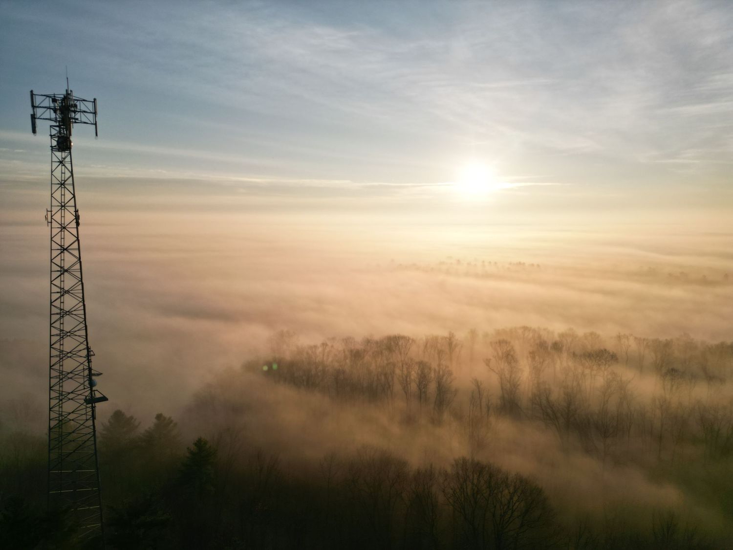 Aerial view of a foggy landscape at sunrise with a tall telecommunications tower on the left and misty treetops below.