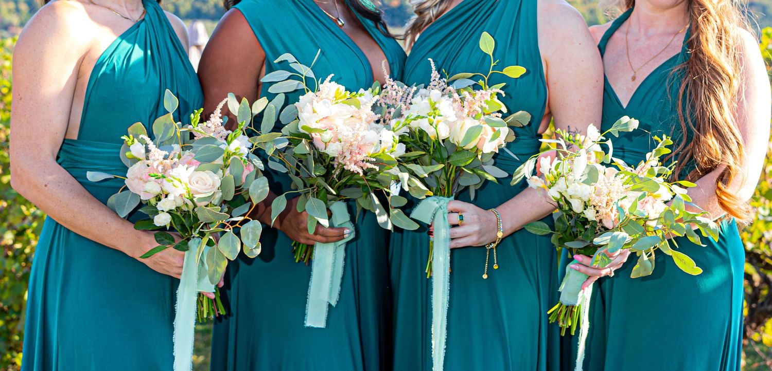 Bridesmaids in teal dresses hold white floral bouquets during outdoor wedding ceremony.