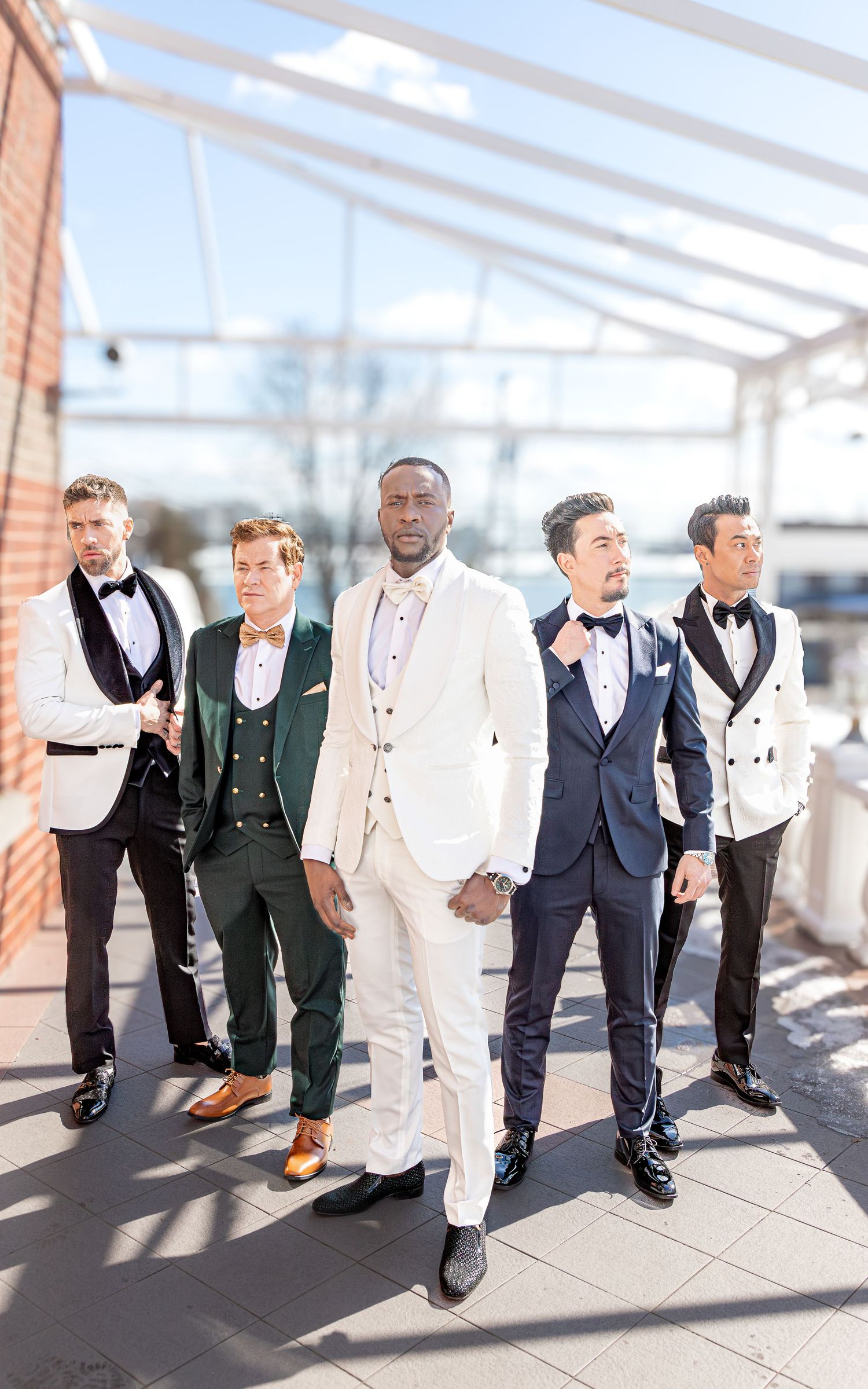 Five groomsmen in coordinated formal attire posing in a modern venue with glass ceiling.