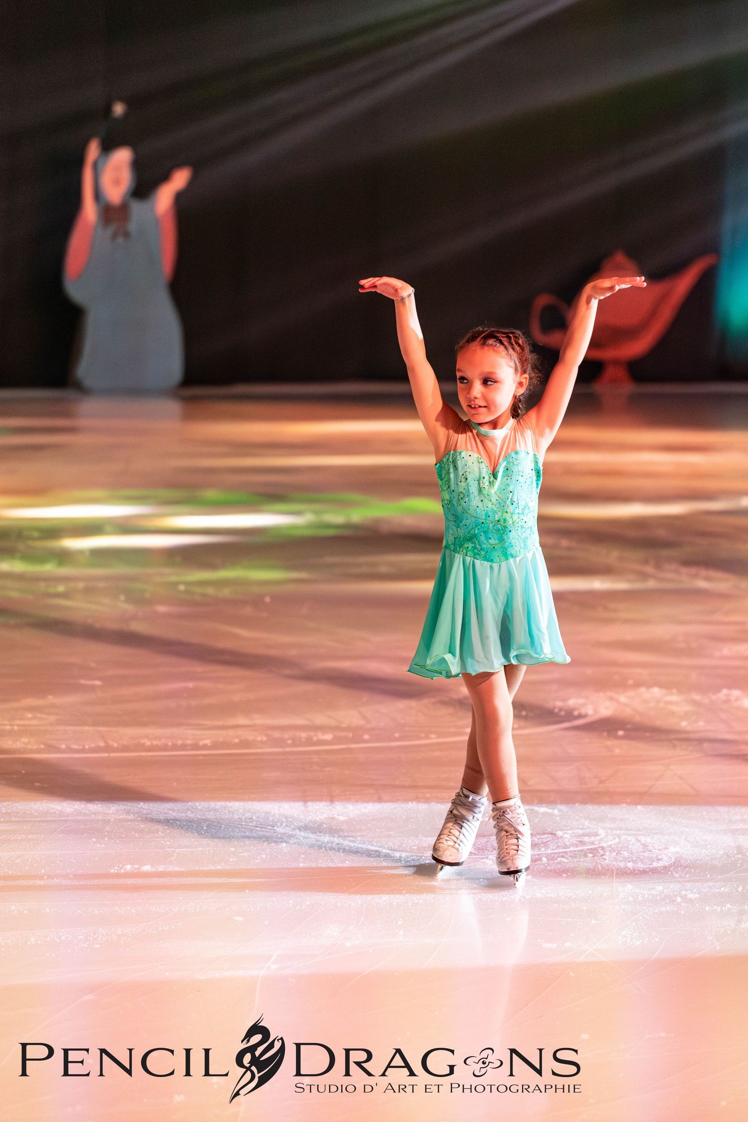 Young girl figure skater performs on ice in a turquoise dress with arms raised during a skating recital.