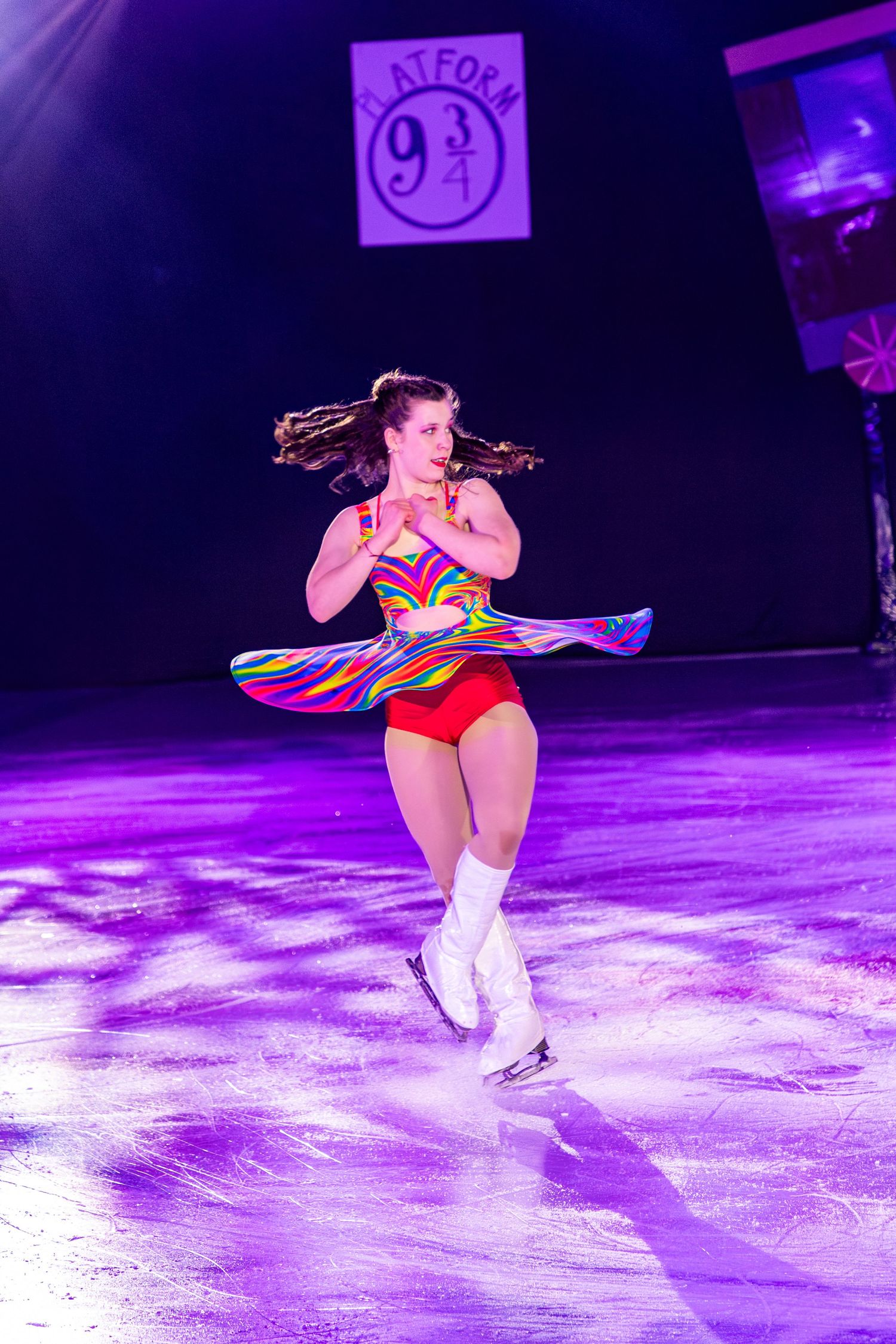 Figure skater in colorful rainbow dress spinning on ice rink with Platform 9 3/4 sign backdrop.