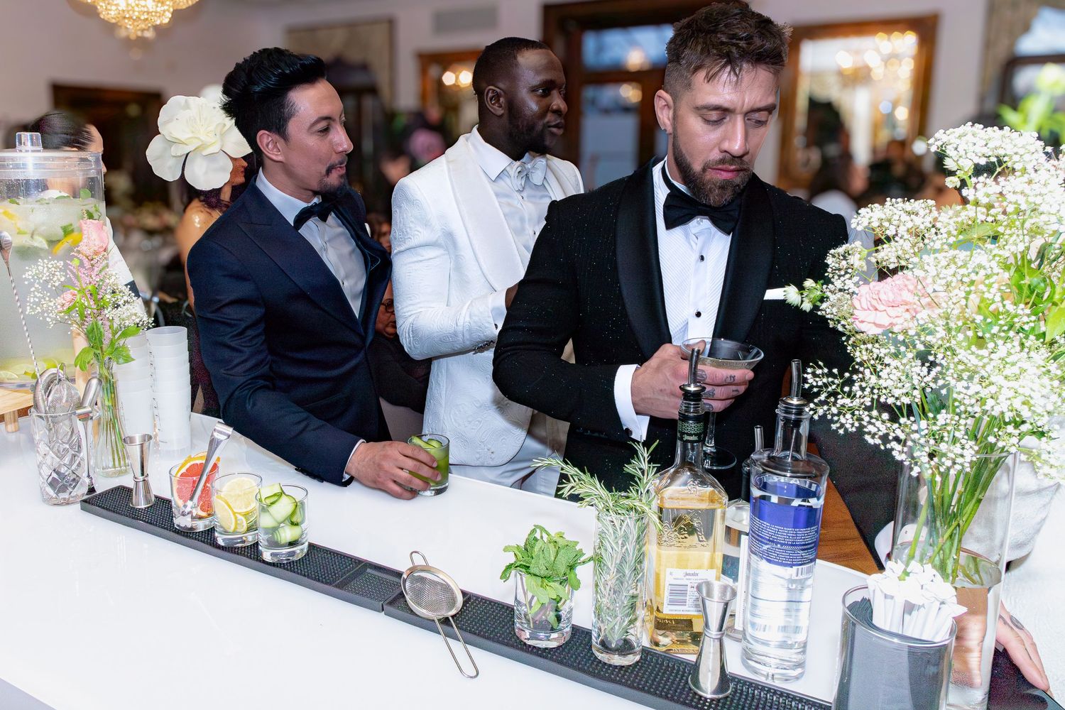 Guests in formal attire gather at an elegant bar decorated with floral arrangements and glassware at an upscale event.