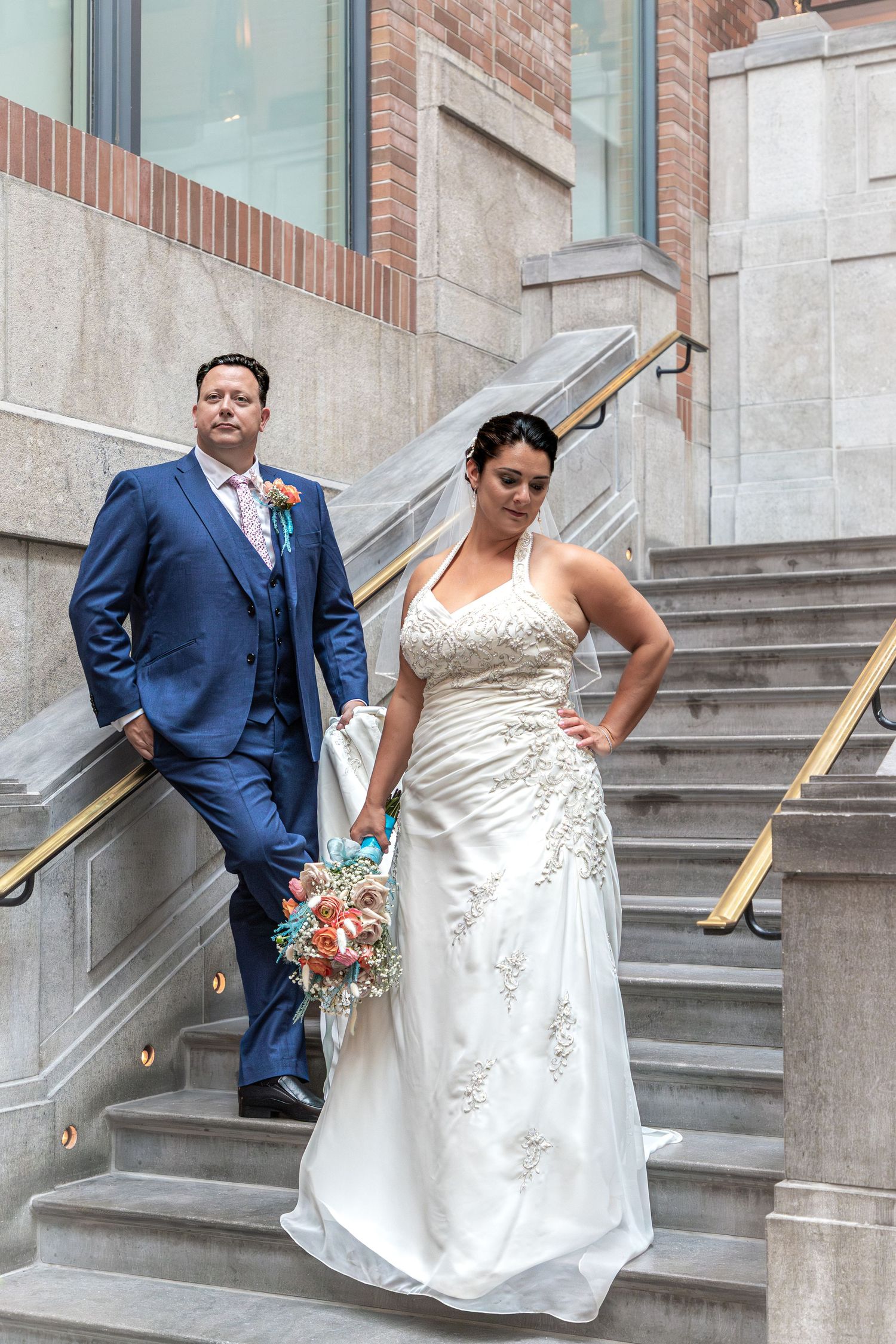 A couple poses on elegant stone stairs, with the bride in a white embellished gown and groom in a navy blue suit.