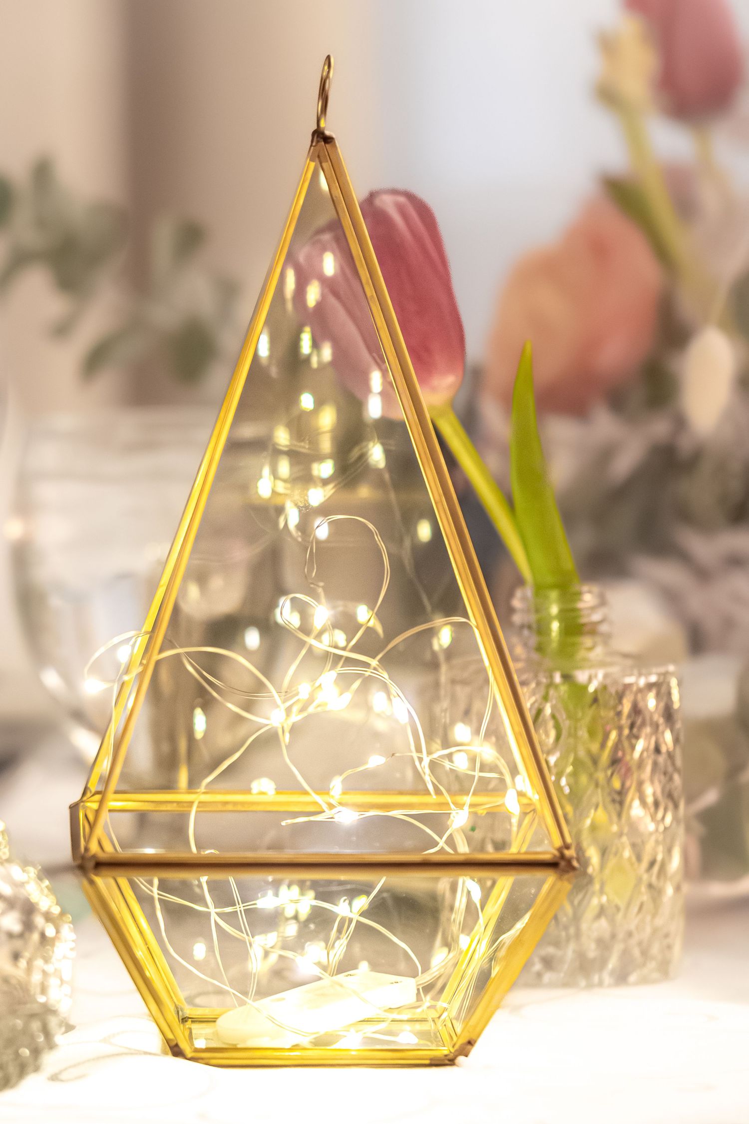 Gold geometric terrariums with delicate fairy lights create a magical table decoration display against a soft blurred background.
