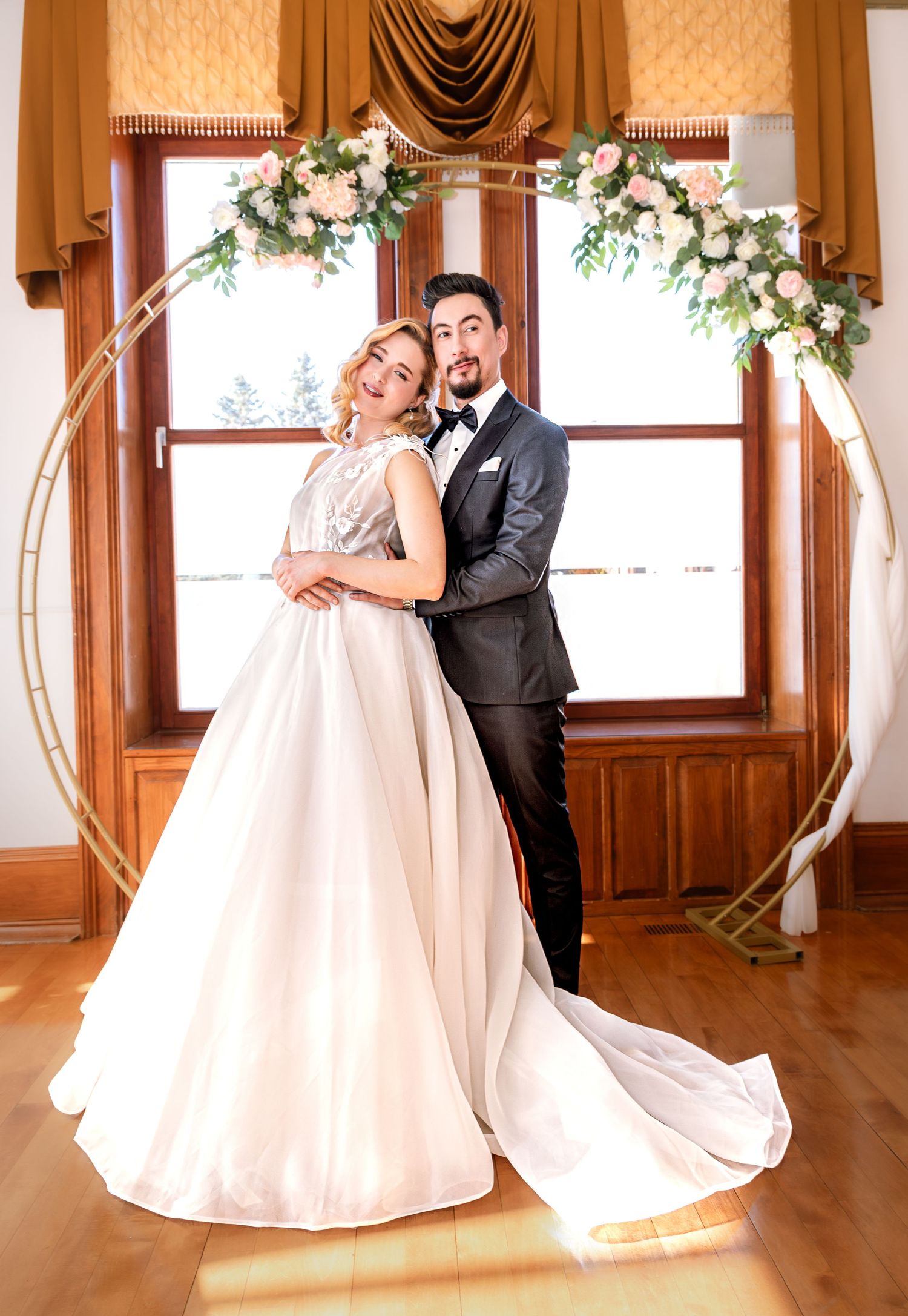 Wedding couple dancing in elegant room with wooden floors and draped windows.