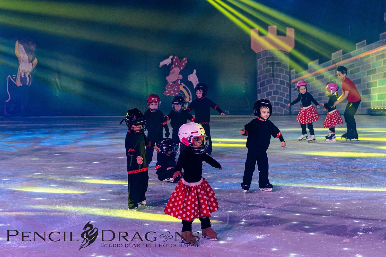 Children in costumes perform an ice skating show on a rink with colorful stage lighting and a castle backdrop.