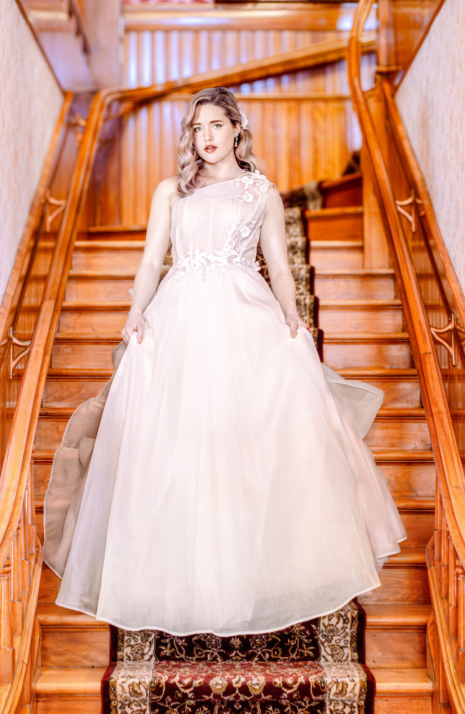 A bride in a white dress stands on an elegant wooden staircase with ornate carpeting and warm lighting.