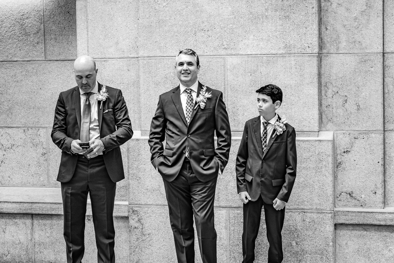 Black and white photo of three generations in formal attire standing against a stone wall at a wedding celebration.
