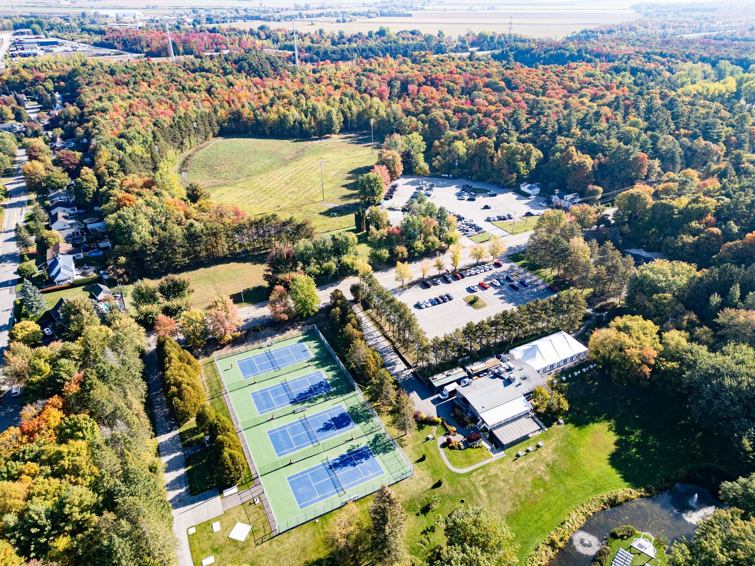 Aerial view of tennis and pickleball courts surrounded by lush autumn foliage and green fields in a suburban setting.