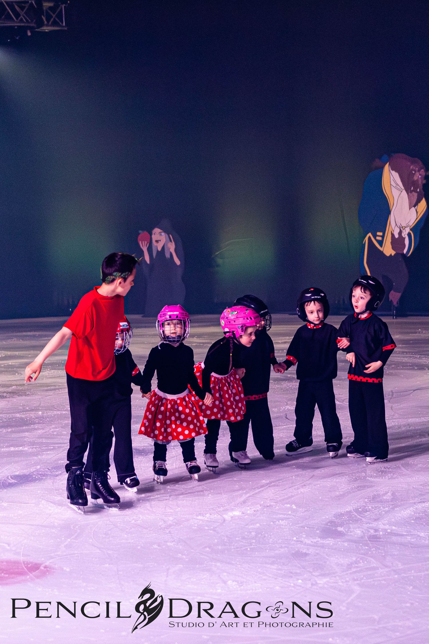 Young children in costumes ice skating with an instructor on a rink, performing in a Disney-themed show.