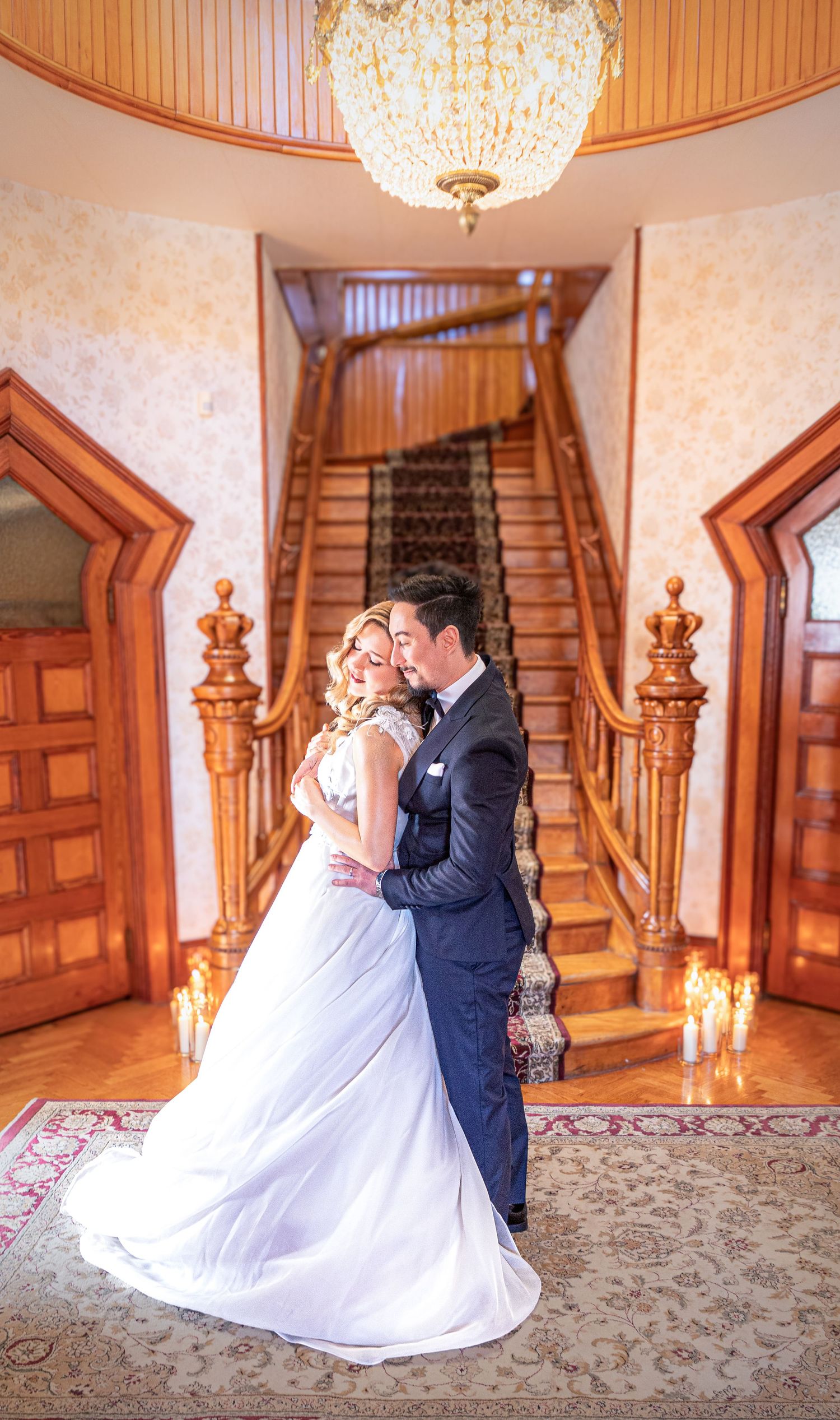 Couple dancing in grand foyer with ornate wooden staircase and crystal chandelier.