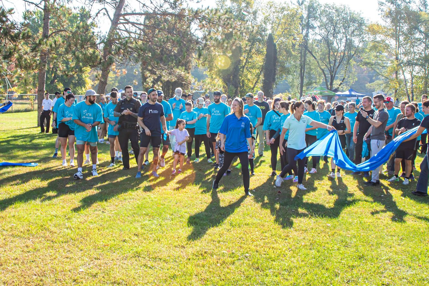 Large group of people wearing teal shirts gathered outdoors in a sunny park for a charity walk event.