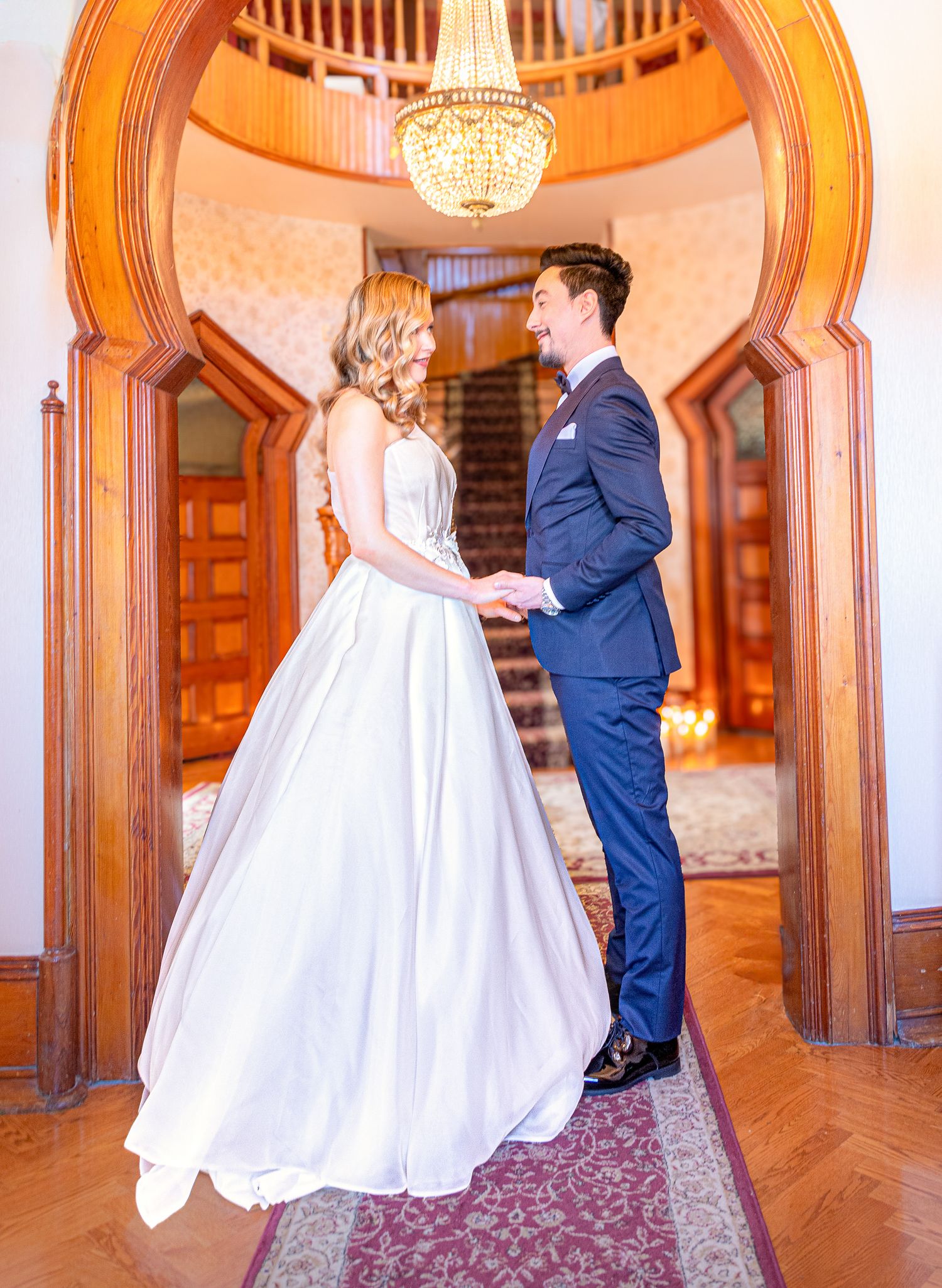 Couple in wedding attire dancing and twirling in an elegant ballroom with ornate woodwork and chandeliers.