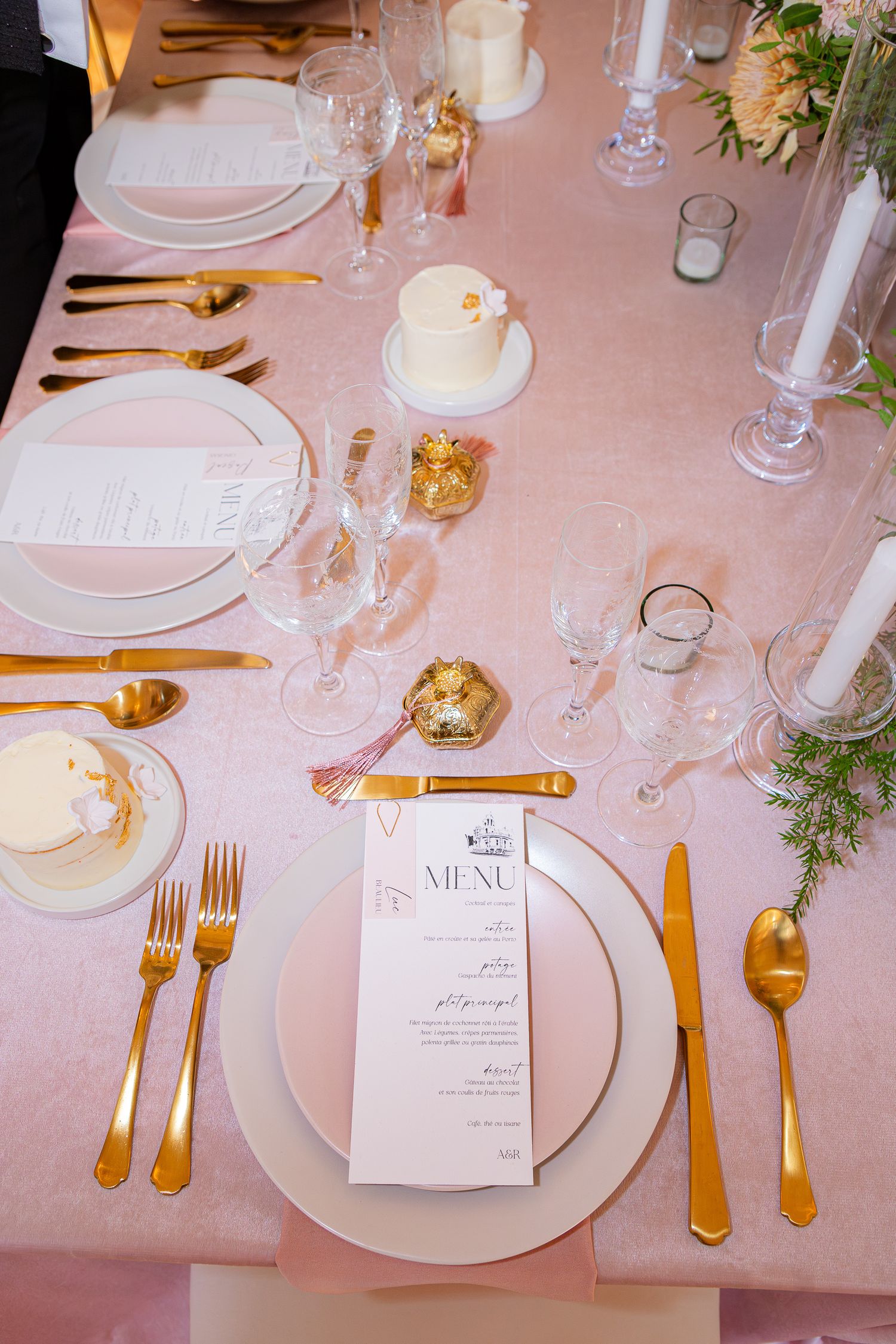 Elegant table settings with gold cutlery, white plates, and menu cards on a soft pink tablecloth for a formal event.